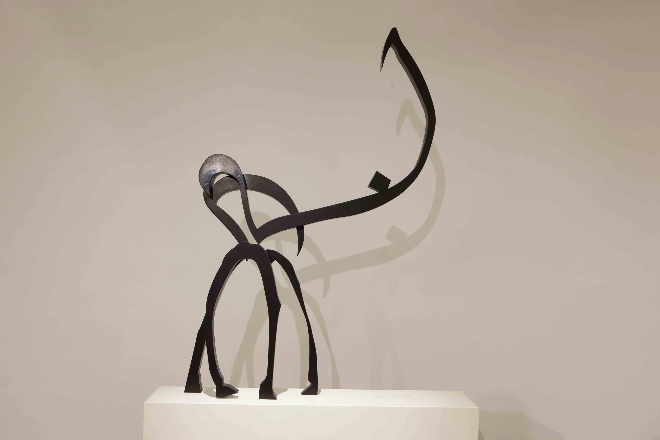 A modern sculpture of a giraffe made from black metal with a large, curved neck and shadow cast on a beige wall.