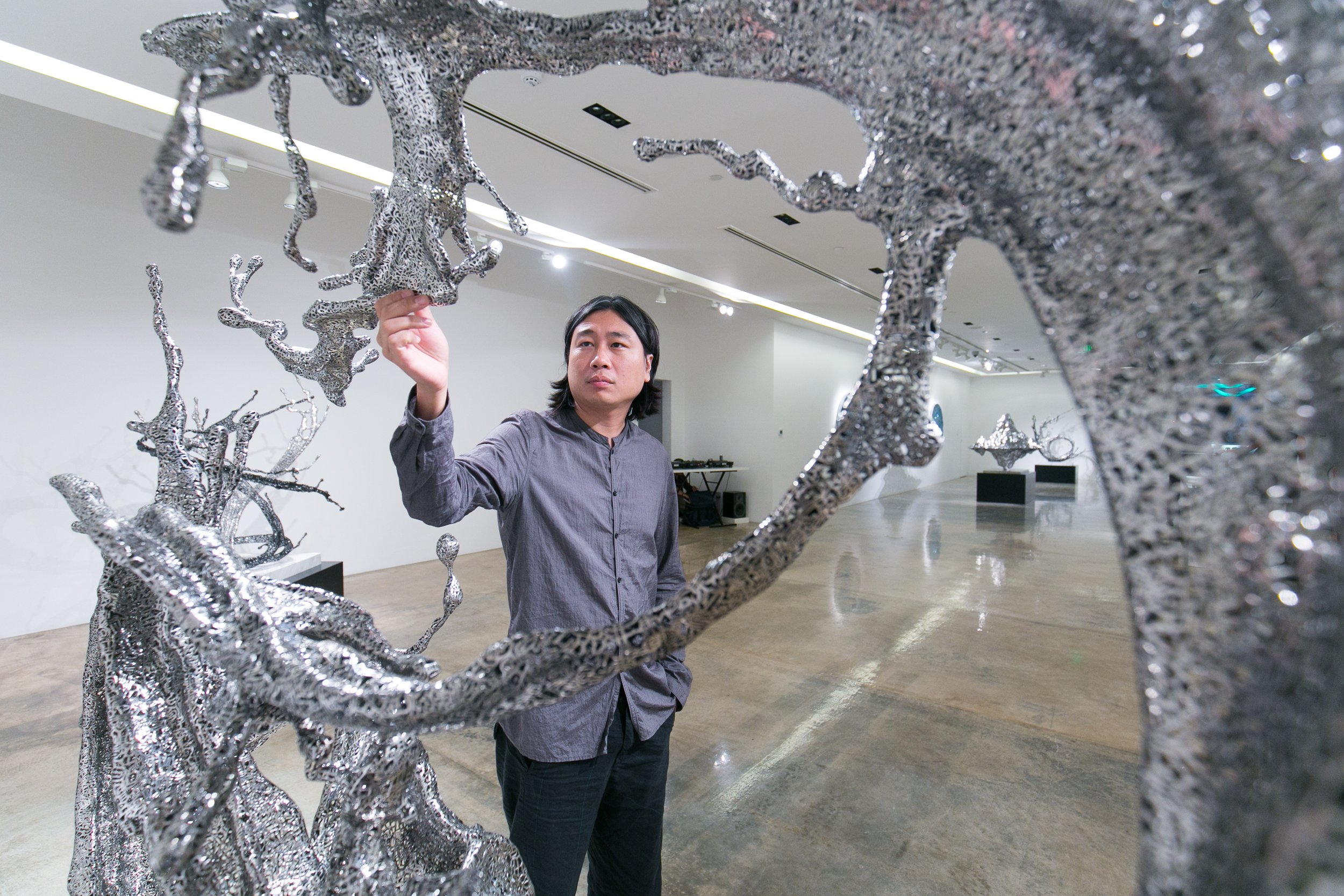 A man in a gray shirt is examining a large, abstract, metallic sculpture in an art gallery. The sculpture has textured, irregular surfaces and is illuminated by gallery lighting.