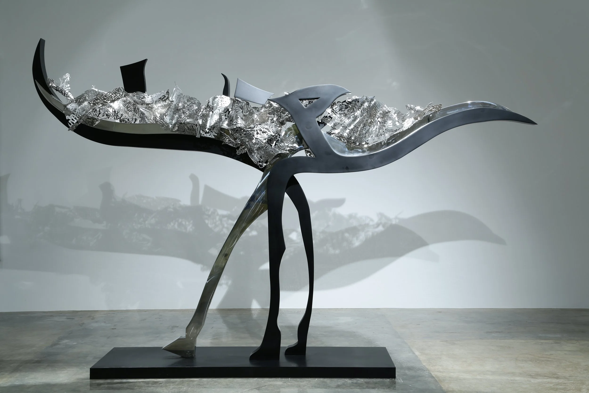 Abstract metal sculpture resembling a stylized bird with elongated wings and legs, casting a shadow on the background.