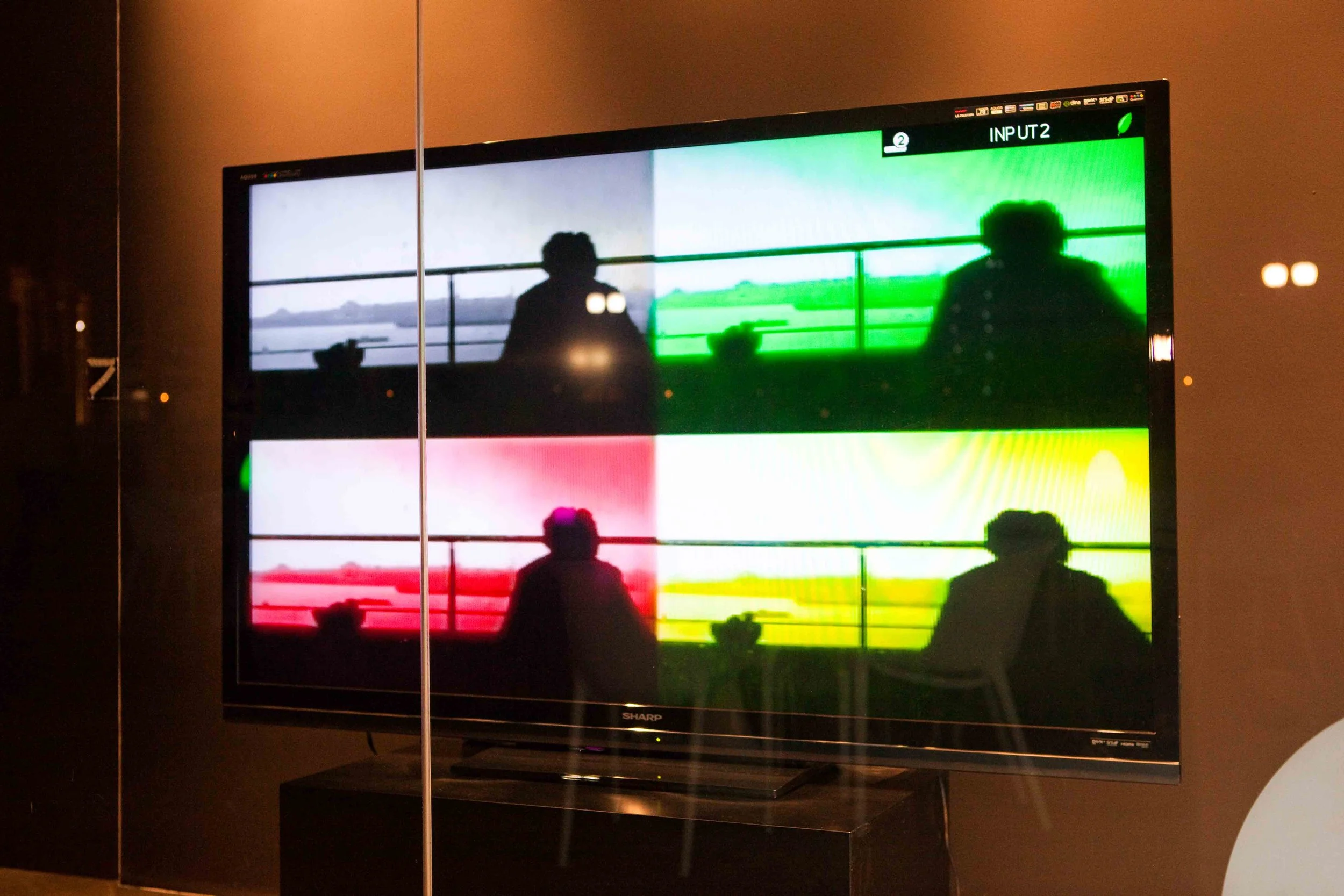 A television screen displaying four separate tinted video feeds of a person sitting on a balcony with a landscape in the background, each feed in a different color filter: black and white, green, red, and yellow.