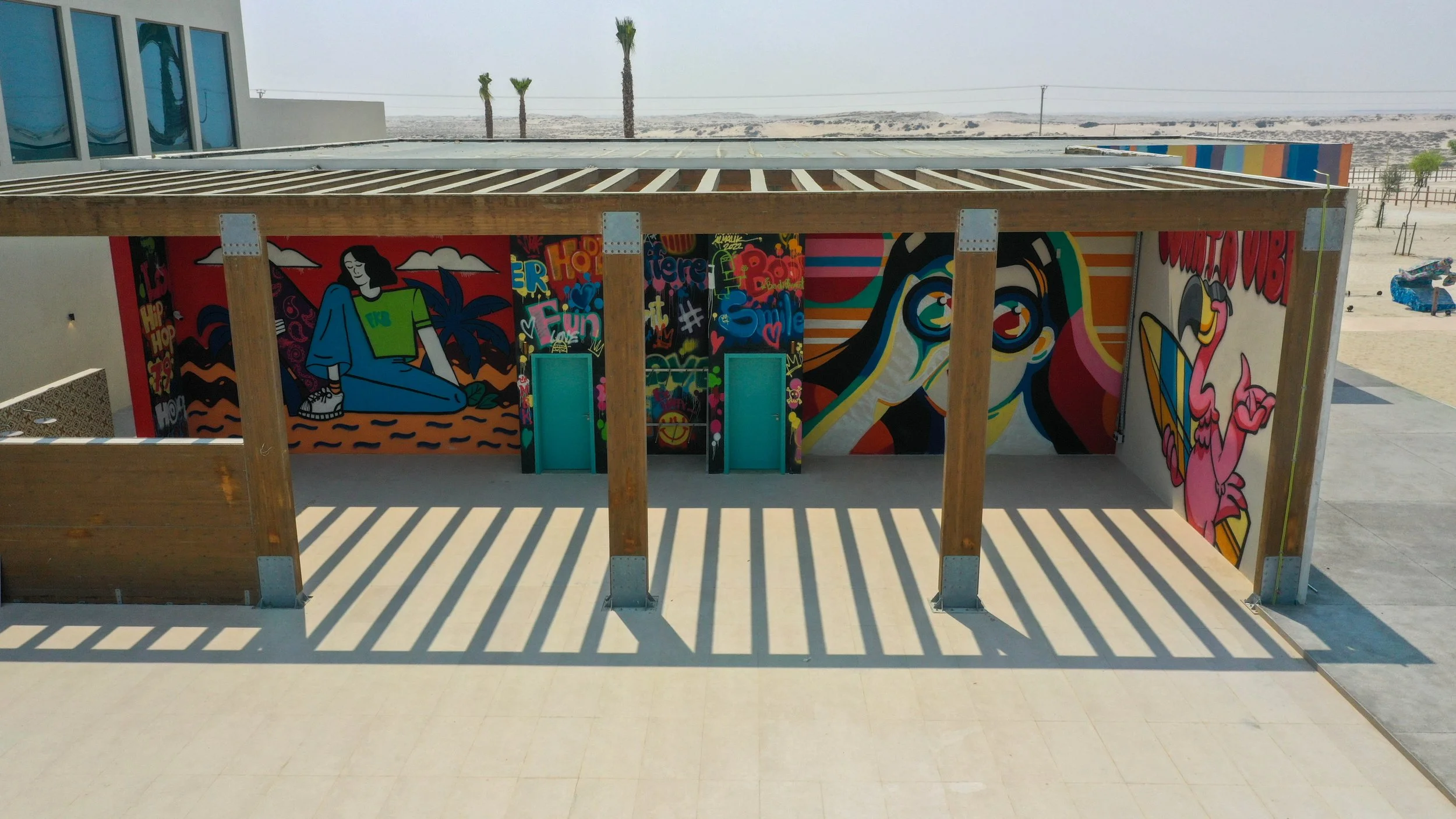 Colorful mural with modern art style depicting a woman, a dog, a flamingo, and various abstract patterns on a wall under a wooden pergola at a beachside location.