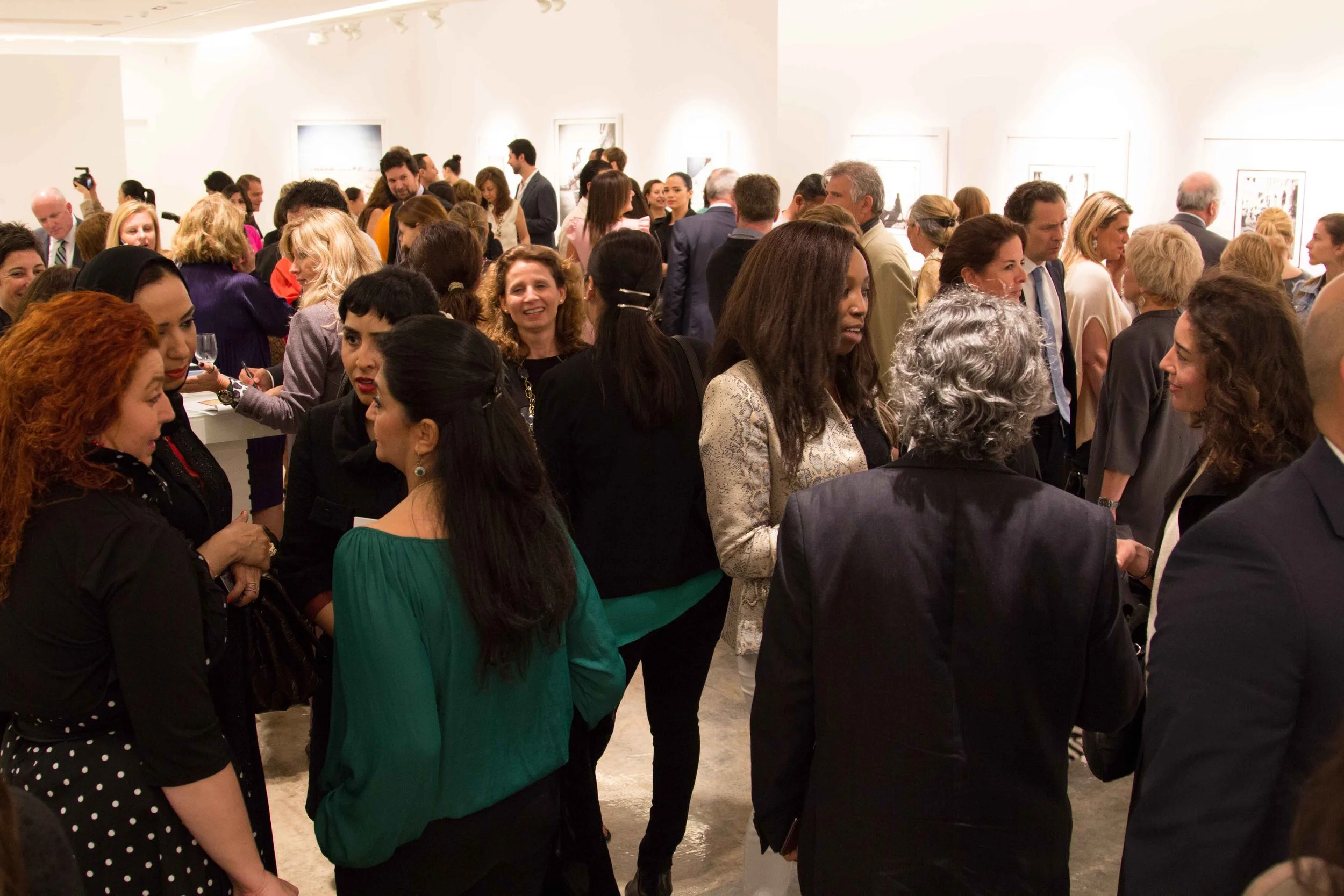 A crowded art gallery with diverse people mingling and viewing artwork on white walls.