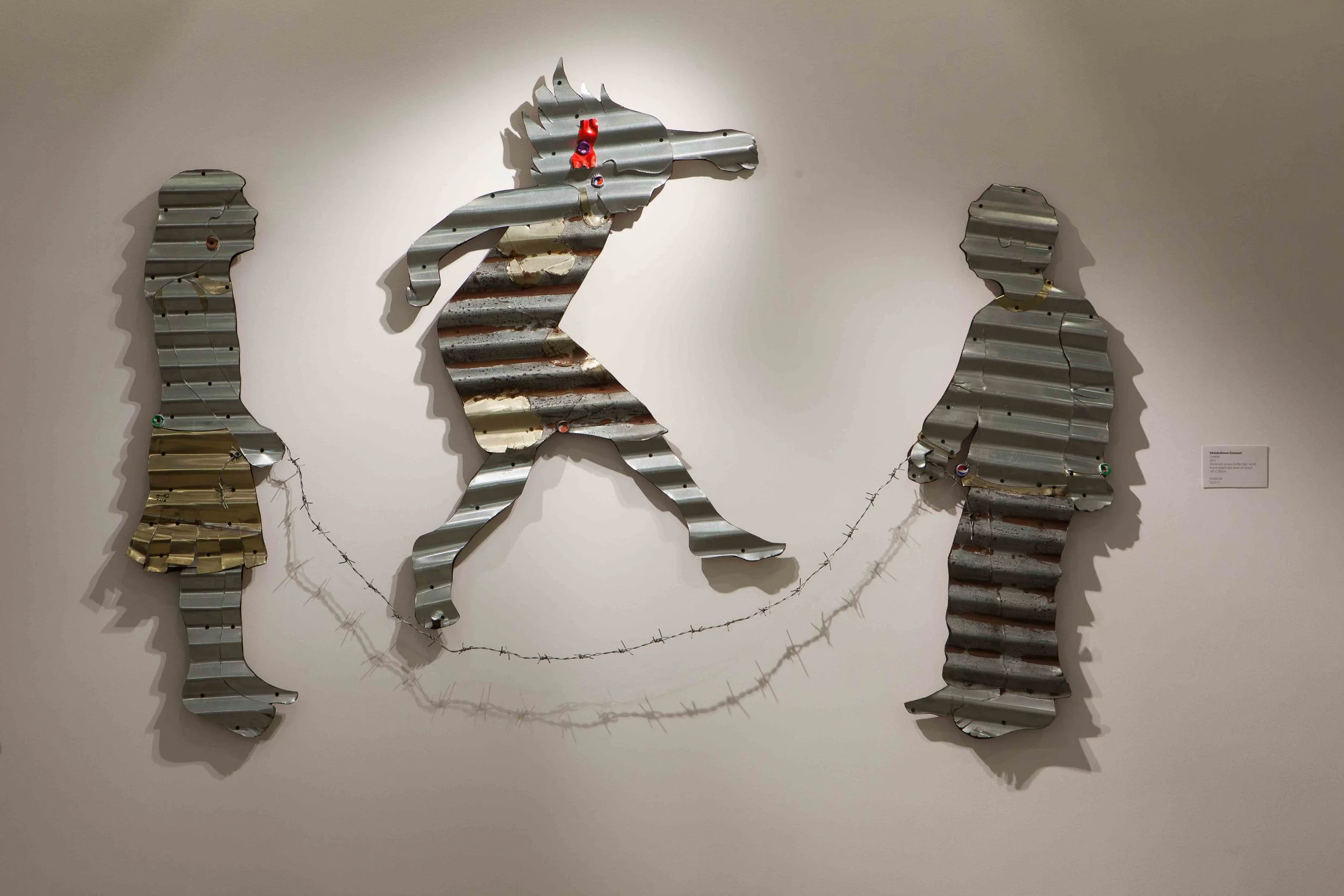 Metal wall art sculpture representing three human figures and a horse, with barbed wire connecting two of the figures, displayed on a white gallery wall.