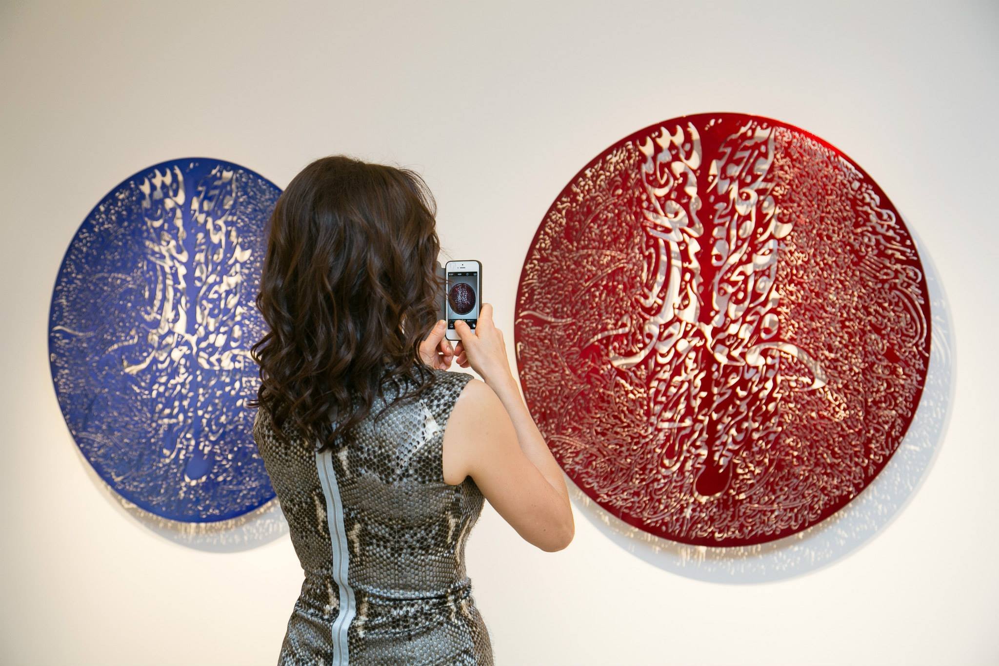 A woman with curly brown hair takes a photo of abstract art, which features a large, circular red design with intricate white and gold patterns, and a similar blue design on the left, on a white gallery wall.