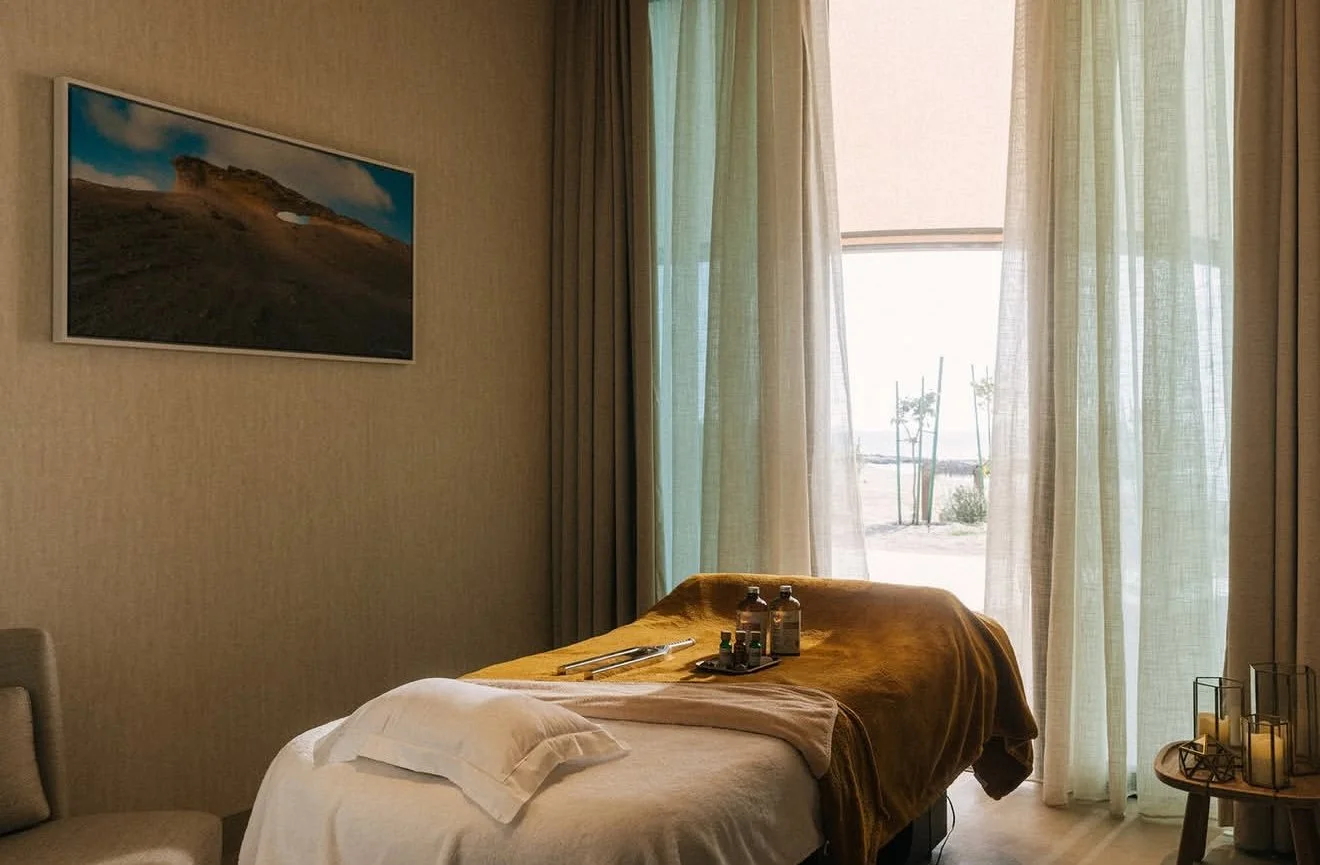 Interior of a spa or massage room with a massage table covered in white and brown towels, bottles of massage oils, and a tray, with a large window revealing a beach scene outside, and a framed landscape picture on the wall.