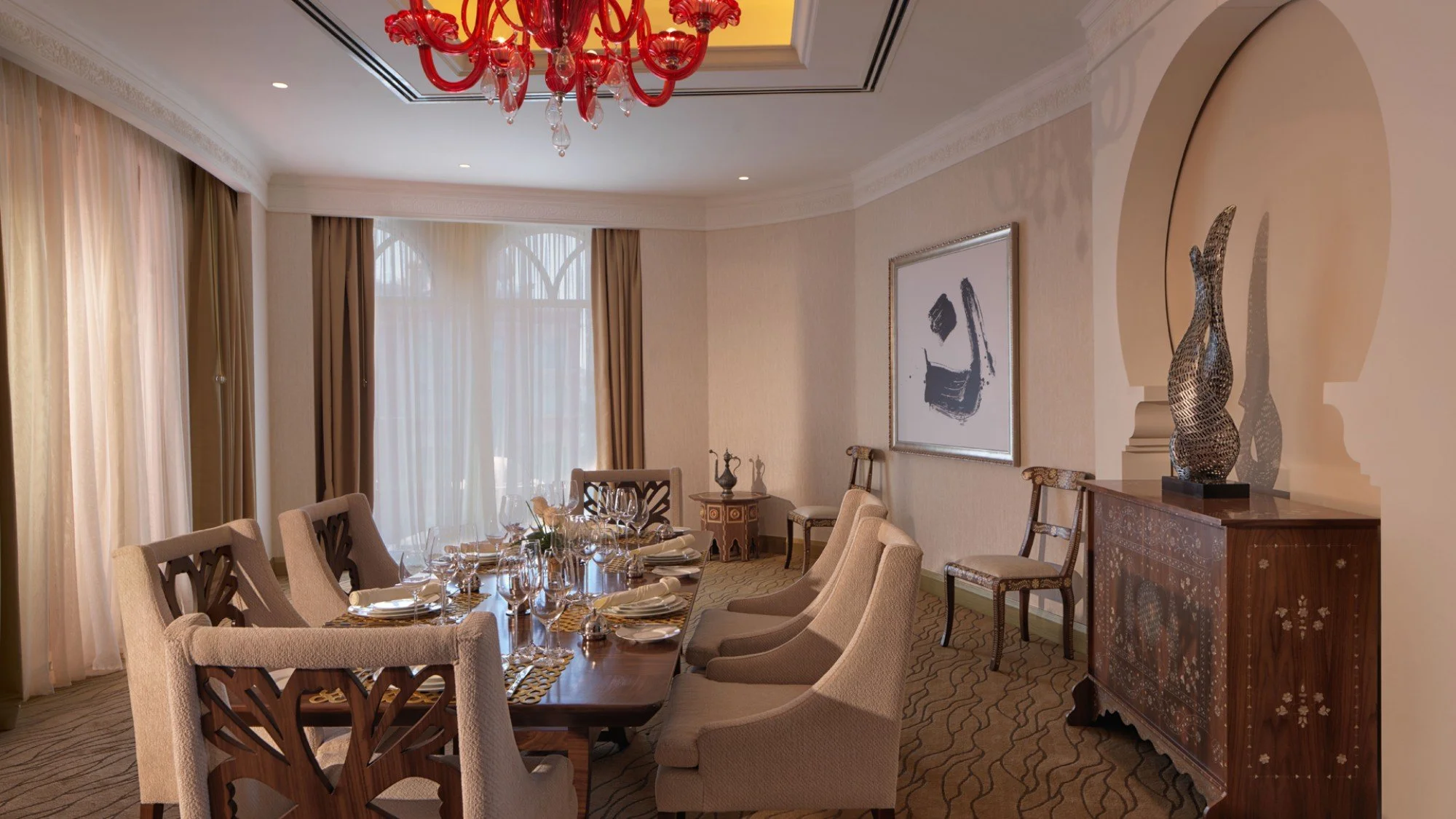 Elegant dining room with a wooden table set for a meal, surrounded by upholstered chairs. Large windows with sheer curtains let in natural light. Decor includes modern artwork, a red chandelier, and a wooden sideboard with decorative vases.