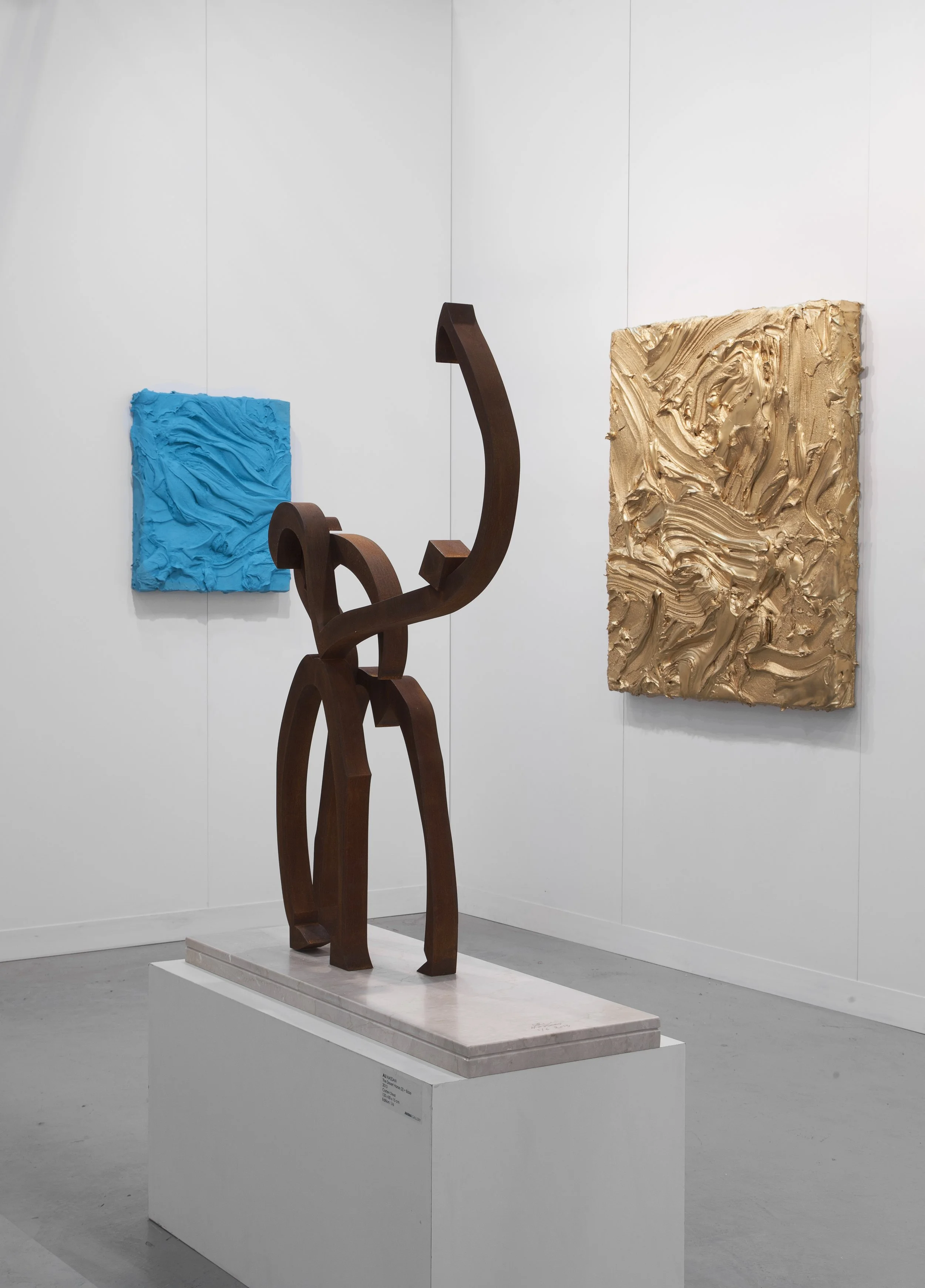 Modern art gallery with abstract sculptures, including a wooden sculpture on a white pedestal and textured paintings on white walls, one blue and one gold.