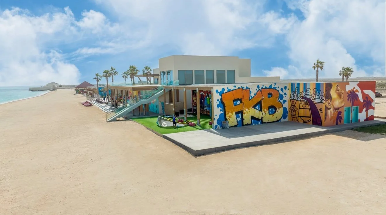 Beachside building with graffiti art, palm trees, and lounge chairs along the sandy shore under a partly cloudy sky.