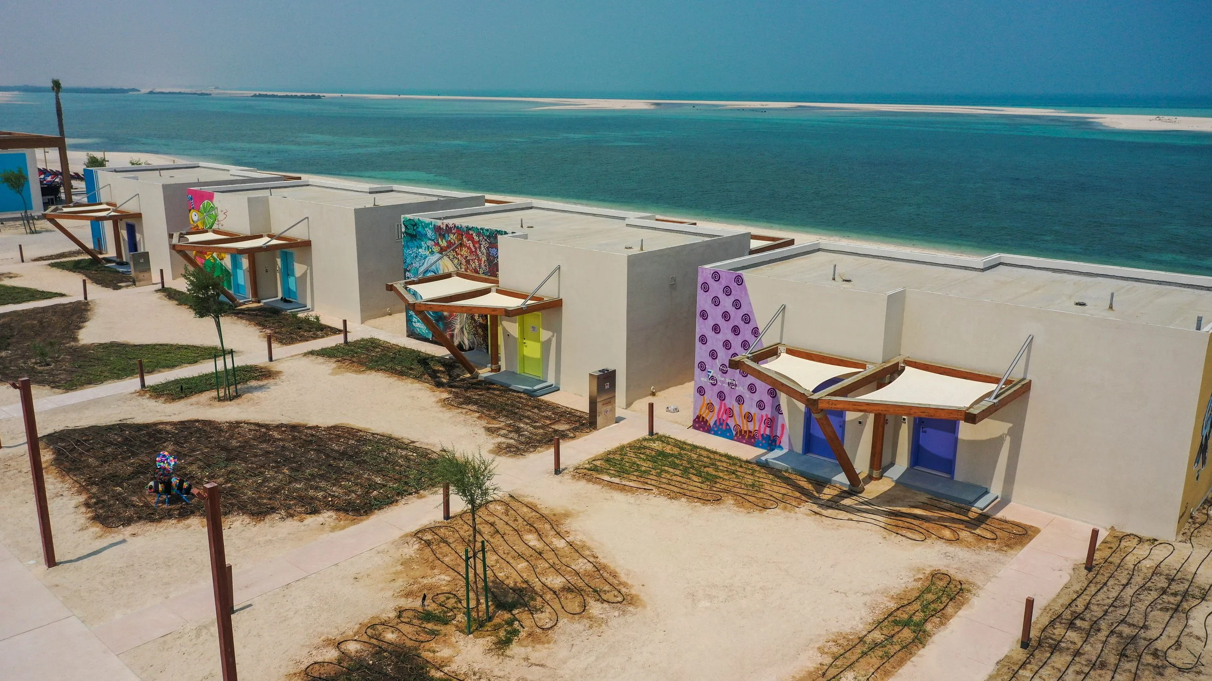 Colorful modern buildings with murals near the beach, surrounded by pathways, small garden plots, and young trees, overlooking the ocean.