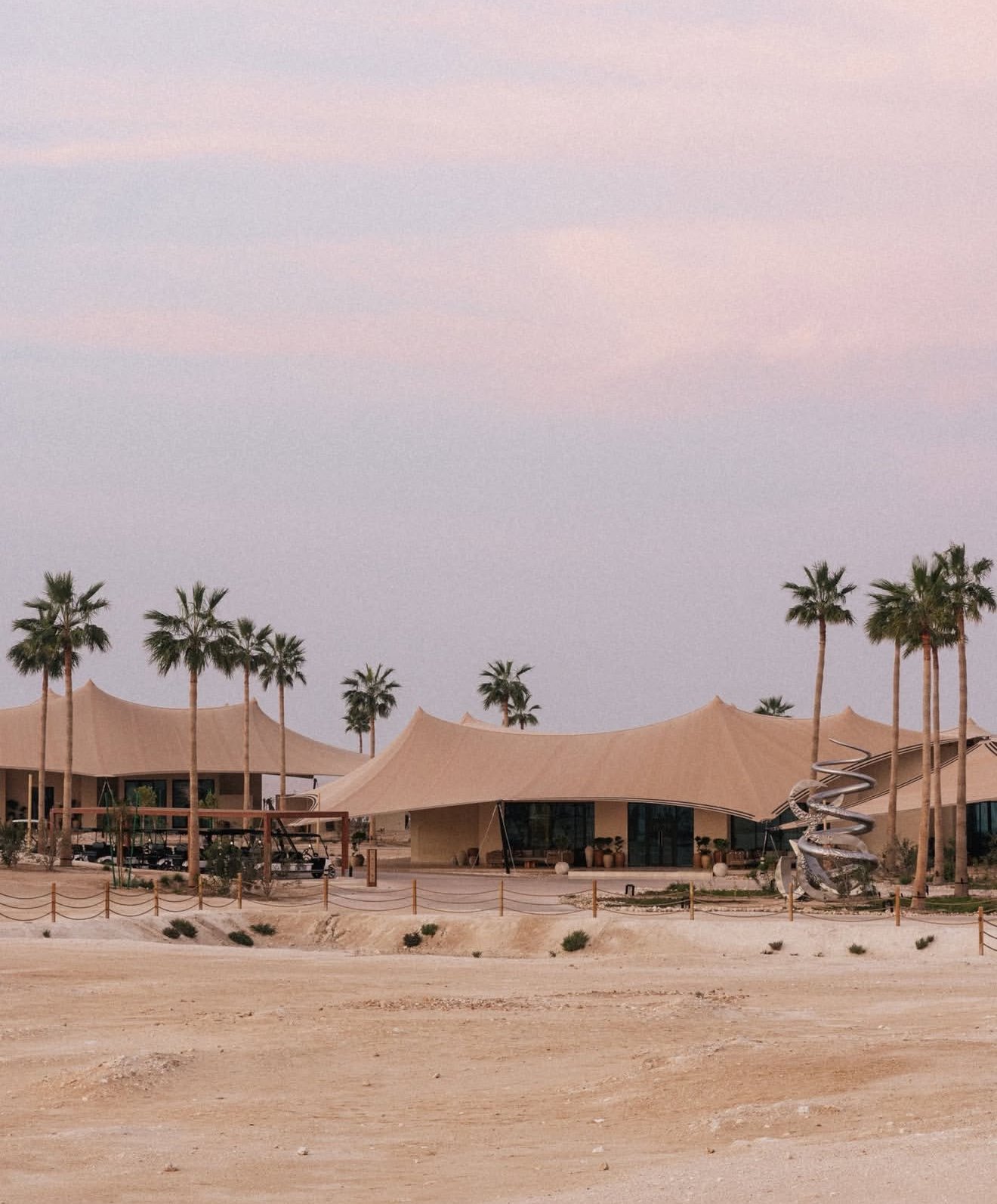 Modern building with fabric roof tents, palm trees, and a spiral slide in a desert landscape at dusk.