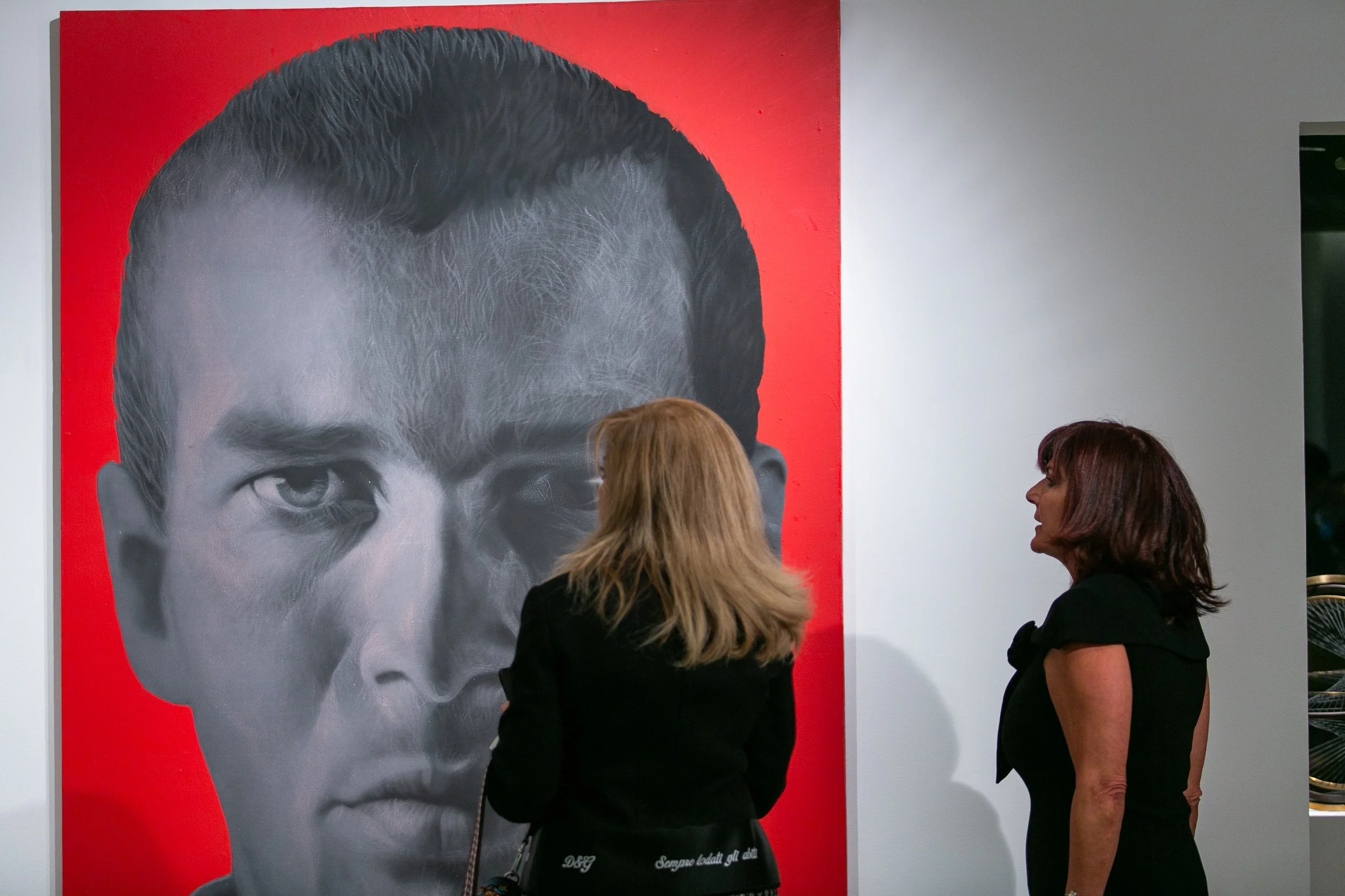 Two women observing a large black-and-white portrait of a man with slicked-back hair on a red background in an art gallery.