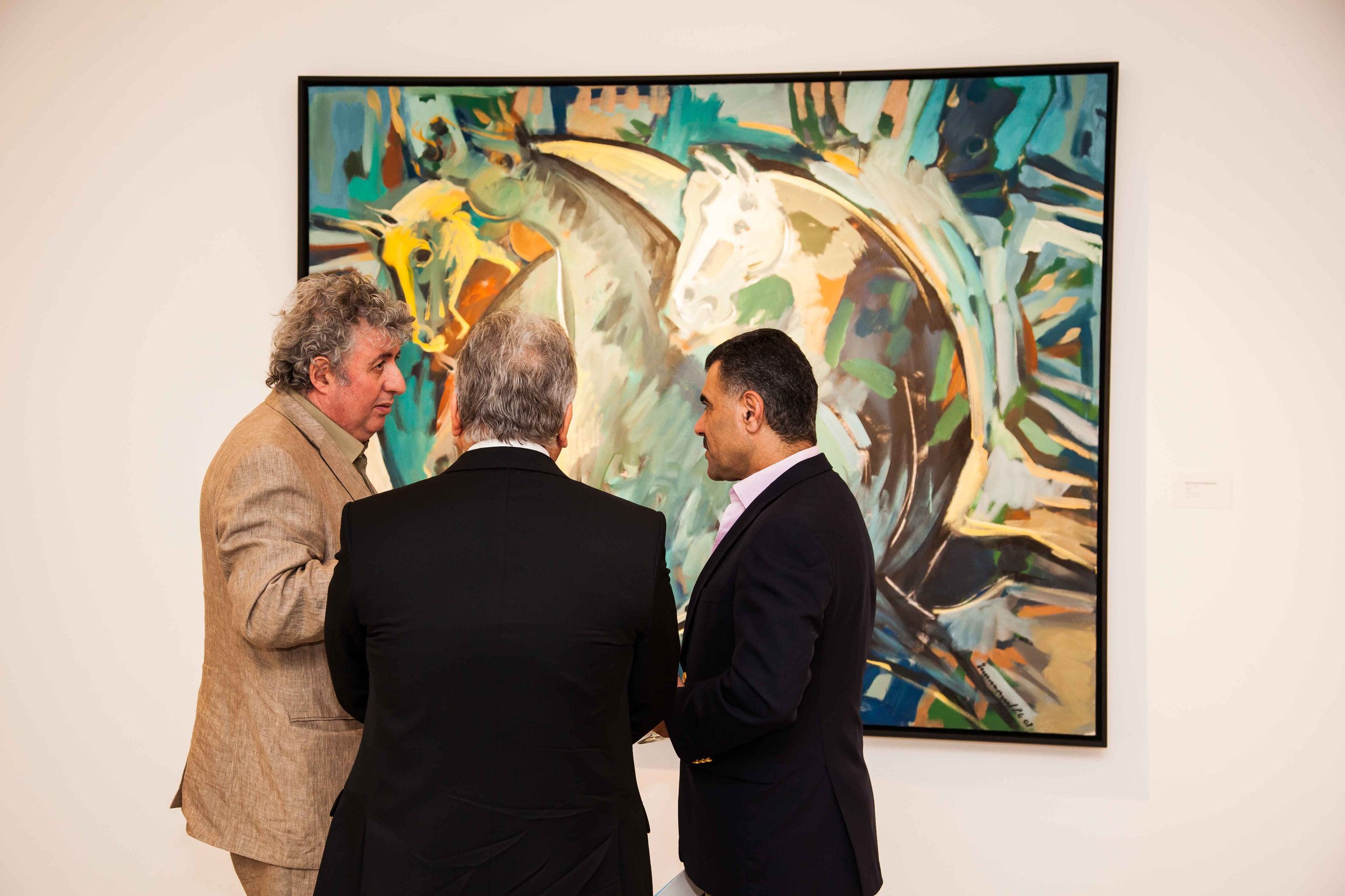 Three men in formal suits talking in front of colorful abstract horse painting at art gallery.