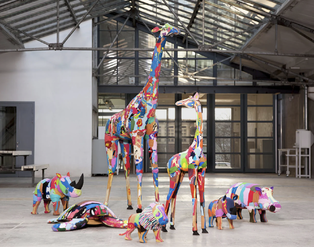 Colorful fabric sculptures of various animals, including a giraffe, zebra, elephant, rhino, and hippopotamus, arranged indoors in a modern, industrial space with metal beams and glass windows.