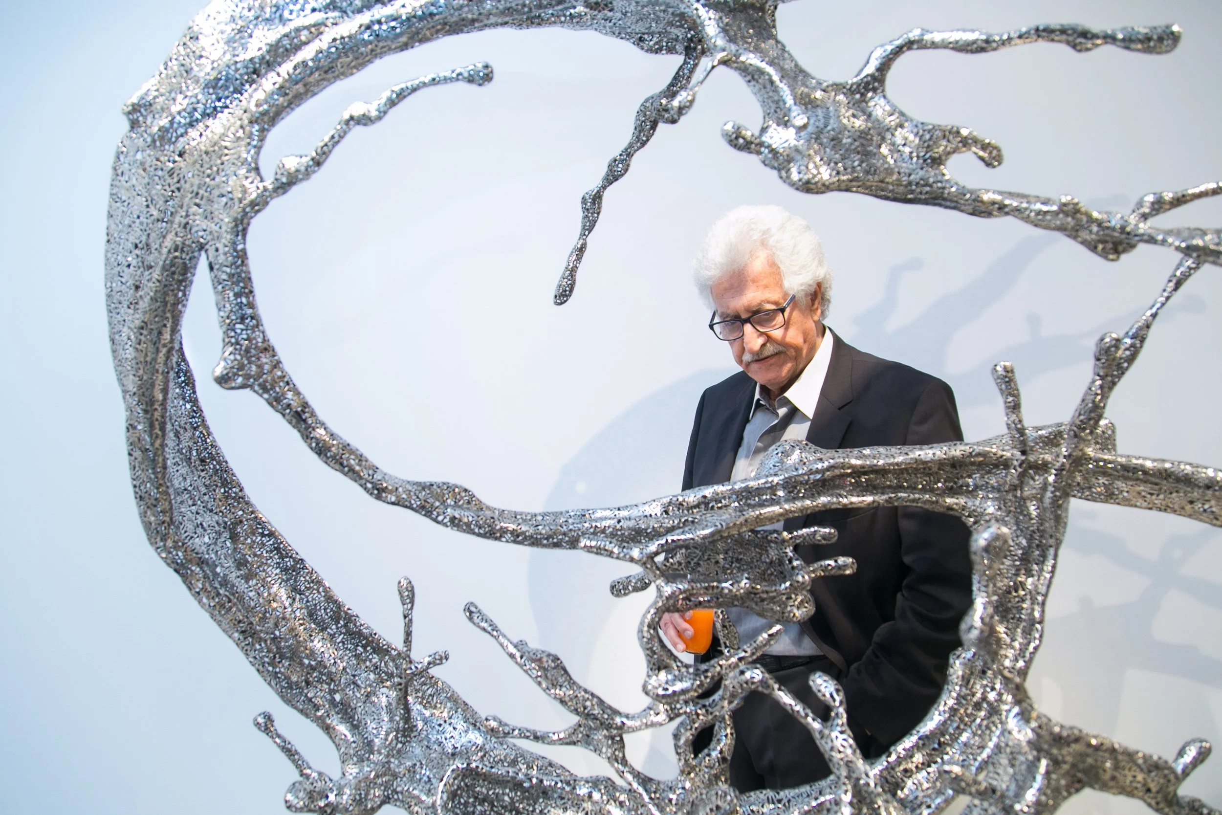 An elderly man with white hair and glasses, dressed in a black suit and gray shirt, looks down while holding an orange-colored object, as he stands behind a modern metallic sculpture resembling intertwined branches forming a circular frame.