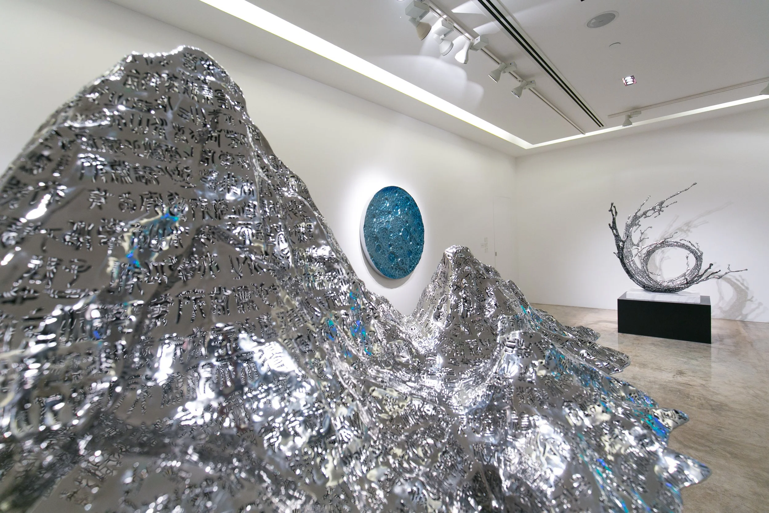 Multiple modern sculptures in an art gallery, including metallic mountain-like structures with Arabic script, a circular textured blue piece on the wall, and an abstract metal sculpture resembling a curled tree branch, all displayed in a well-lit, mi
