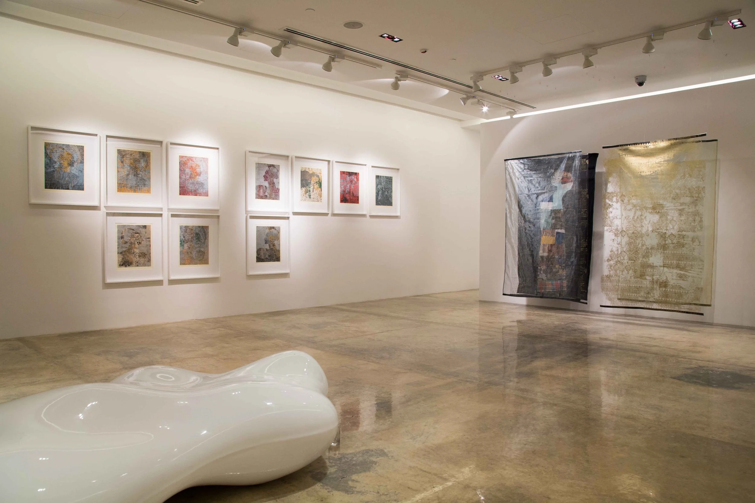 Empty art gallery with framed abstract paintings on the left wall and two large textile artworks on the right wall, a white sculptural bench in the foreground, and polished concrete floors.