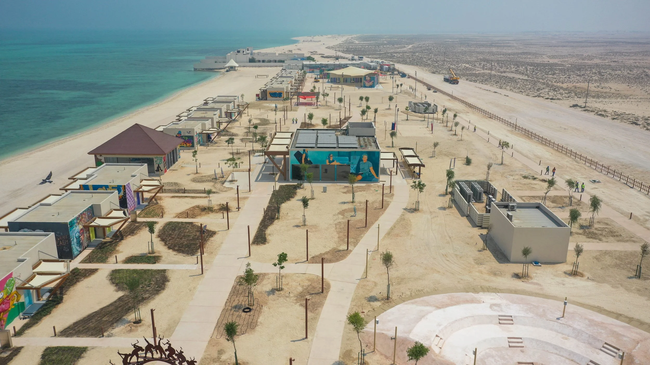 Aerial view of a beachside development with small buildings, colorful murals, pathways, and sparse trees, next to turquoise water and sandy dunes.