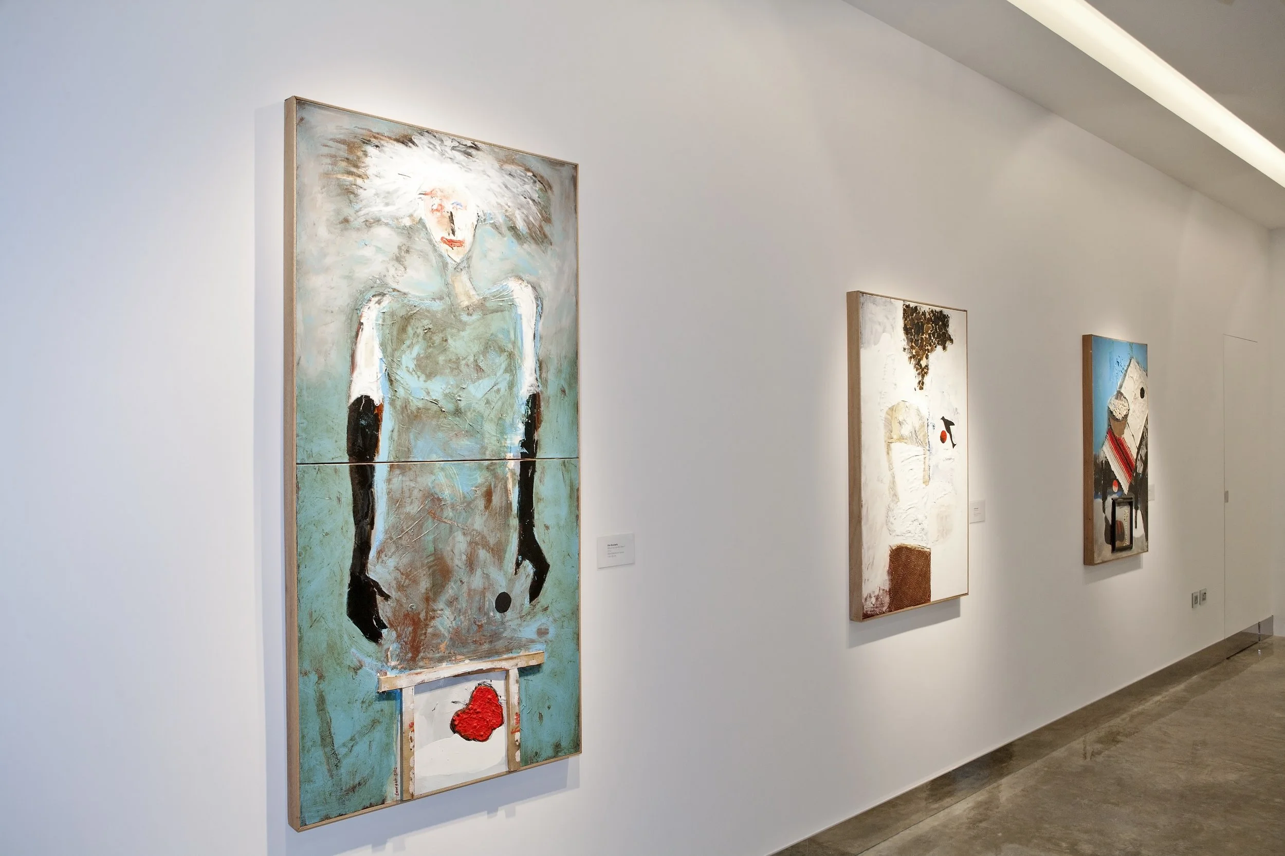 An art gallery display with three abstract paintings on a white wall, showing a woman, a man, and a house, each with textured and colorful brushstrokes.