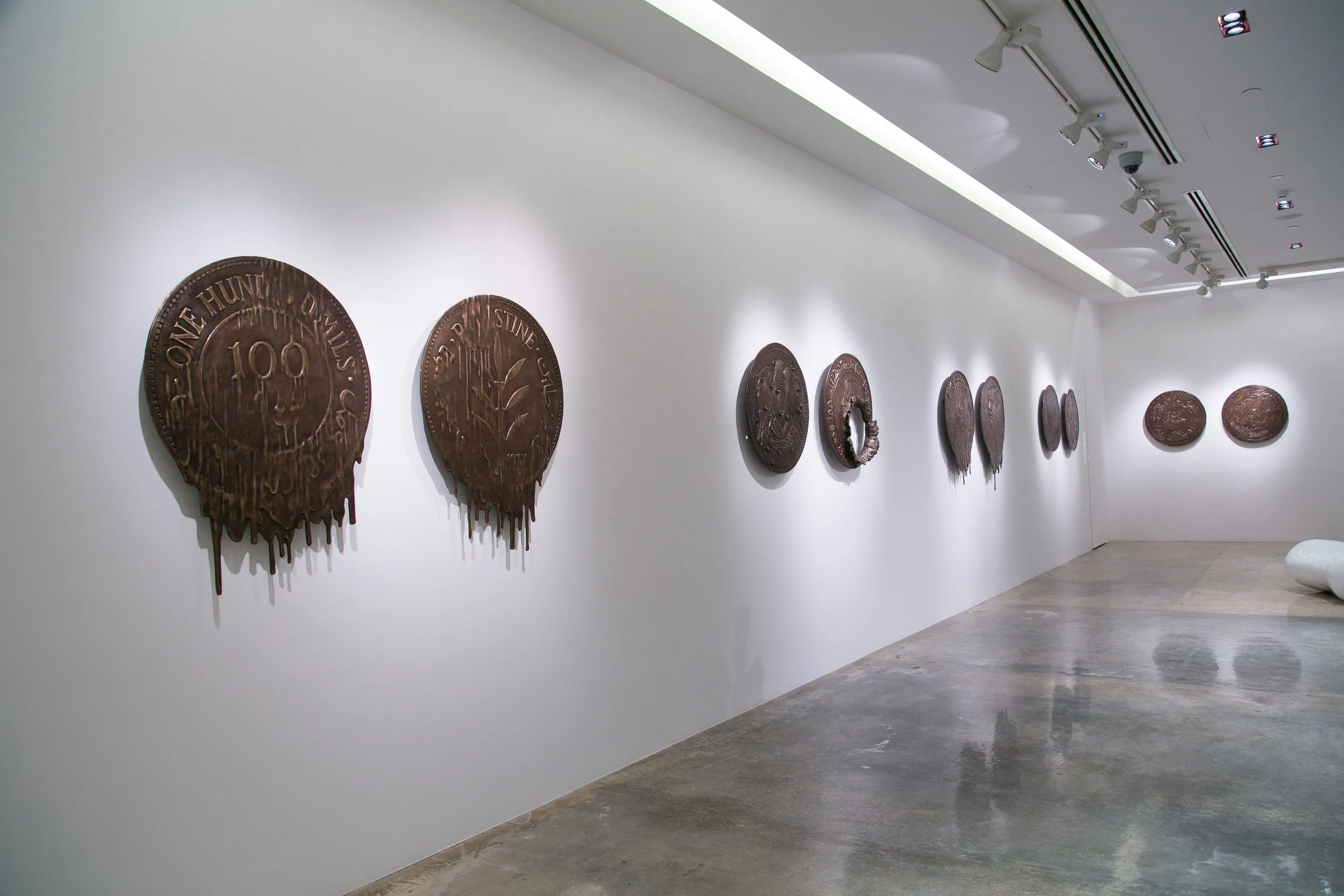 Art installation of multiple bronze medals mounted on a white gallery wall, varying in size and design, with spotlights illuminating each piece.