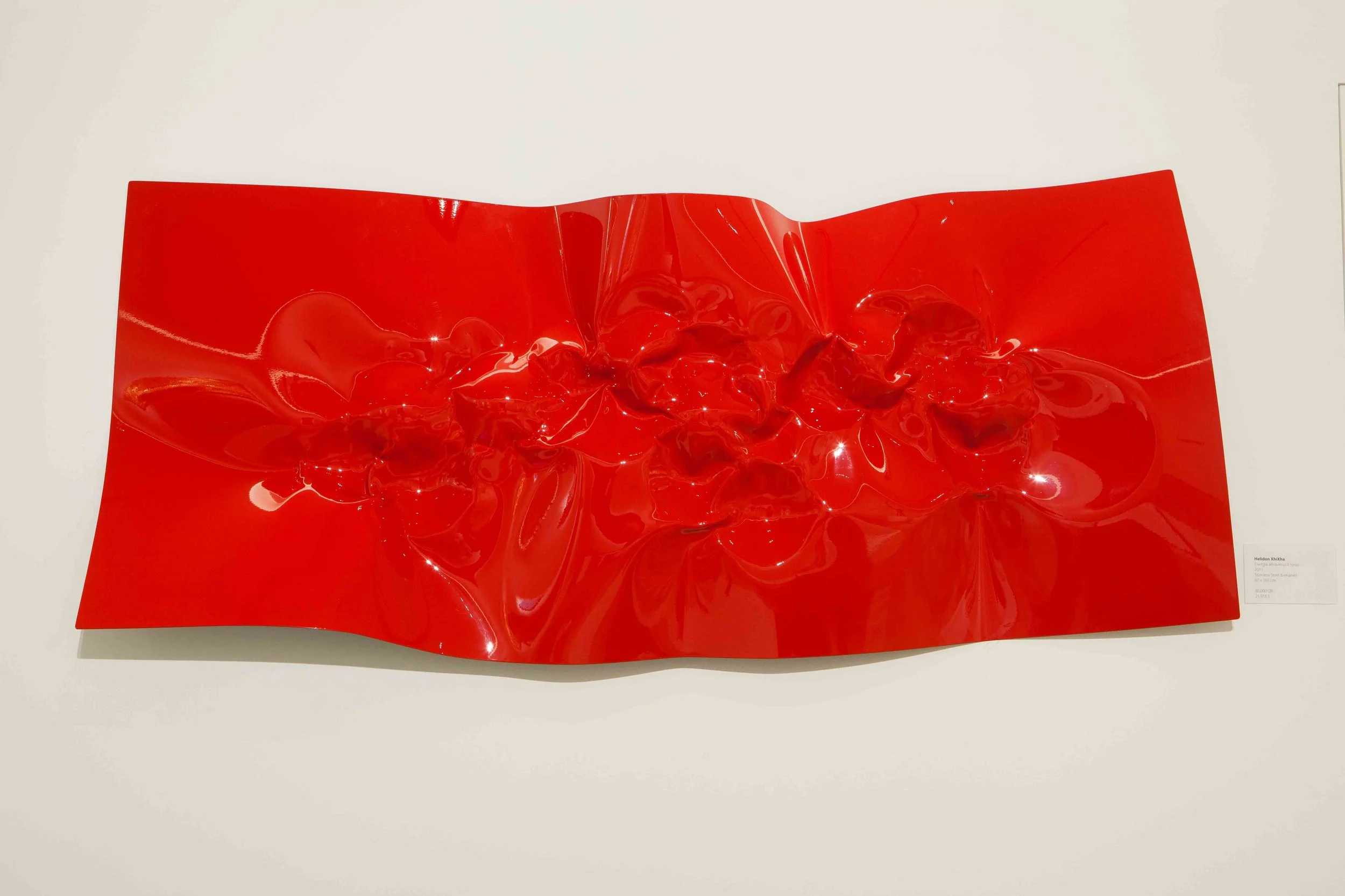 An abstract red, shiny, crumpled sculpture or artwork on a white background.