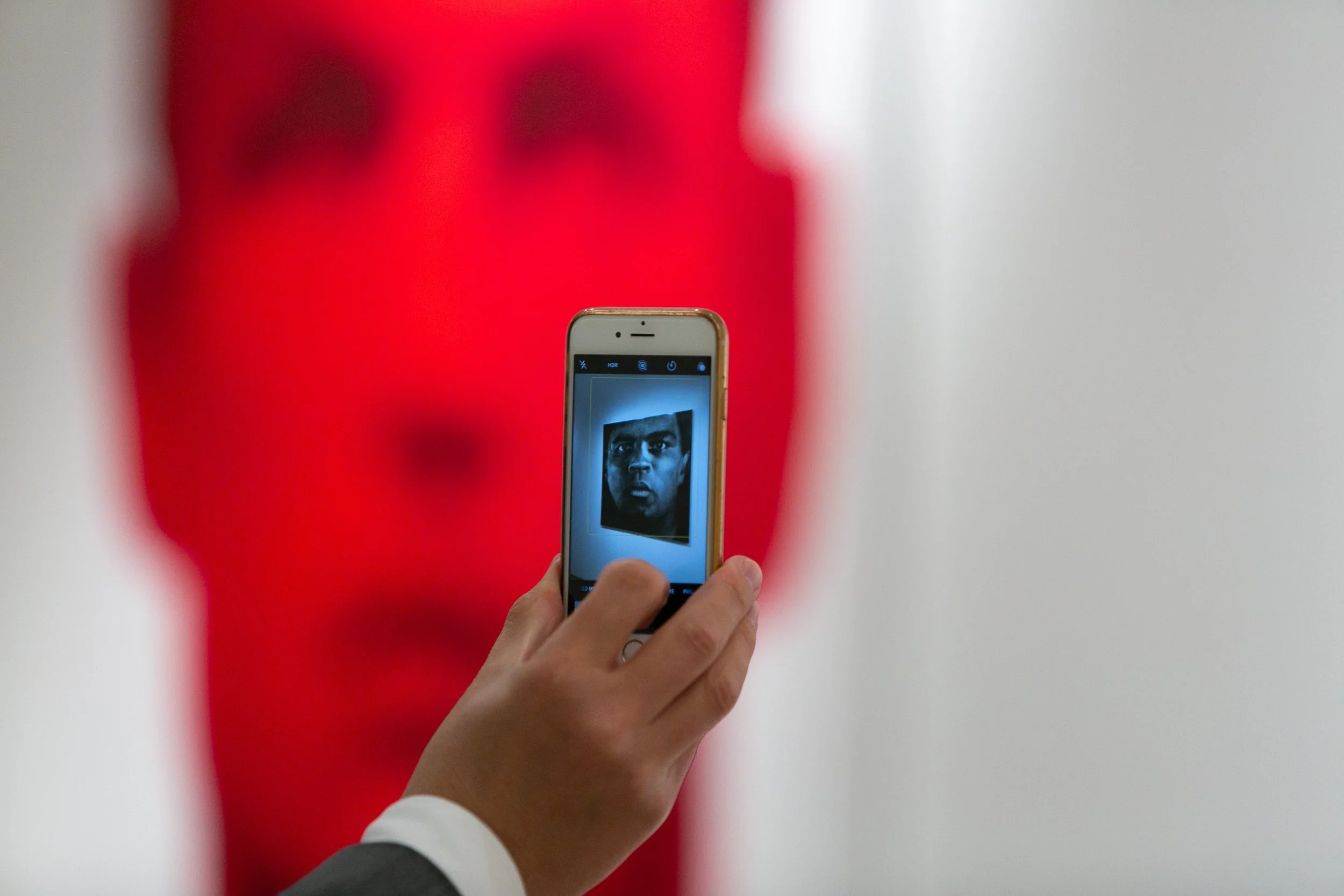 Person taking a photo of a red sculpture or artwork with a smartphone, showing the sculpture's face reflected on the screen.