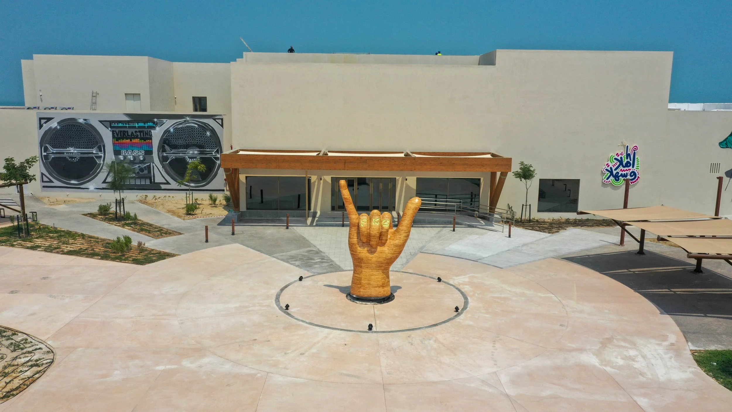 A large outdoor area with a big wooden hand sculpture making the 'rock on' gesture in the center. The area has pathways, small trees, and some shaded seating. A building with a mural of two large boomboxes and graffiti art is in the background, under