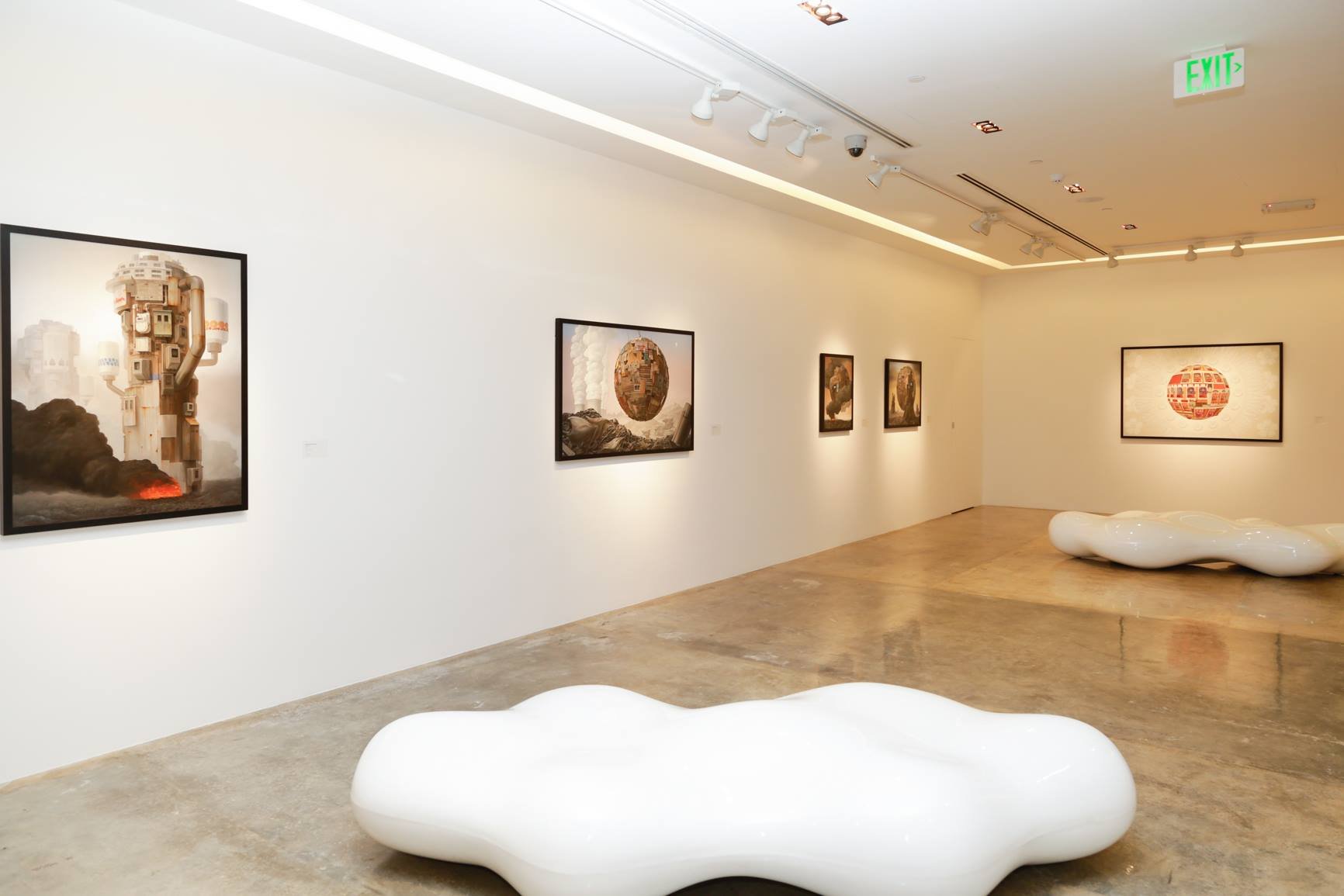 Art gallery with framed artworks on white walls, marble flooring, and white lounge sculptures on the floor, illuminated by track lighting.