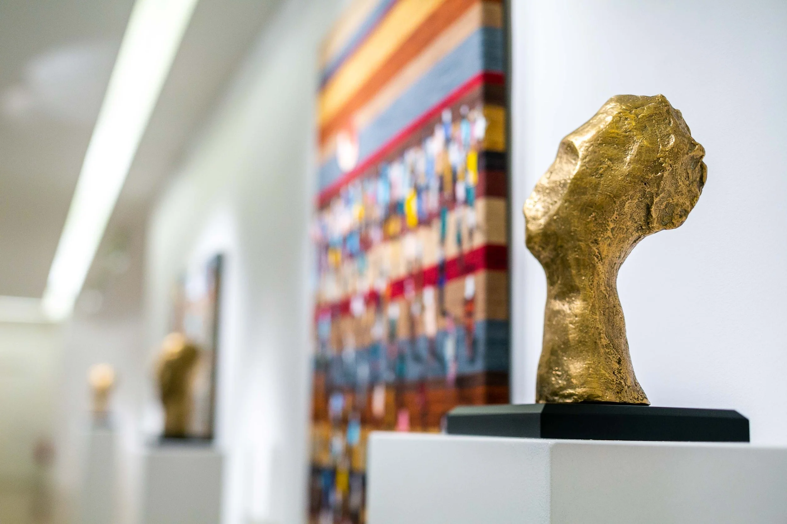 A gold-colored abstract sculpture on a black base displayed on a white pedestal in an art gallery, with a blurred colorful textile artwork on the wall behind it.