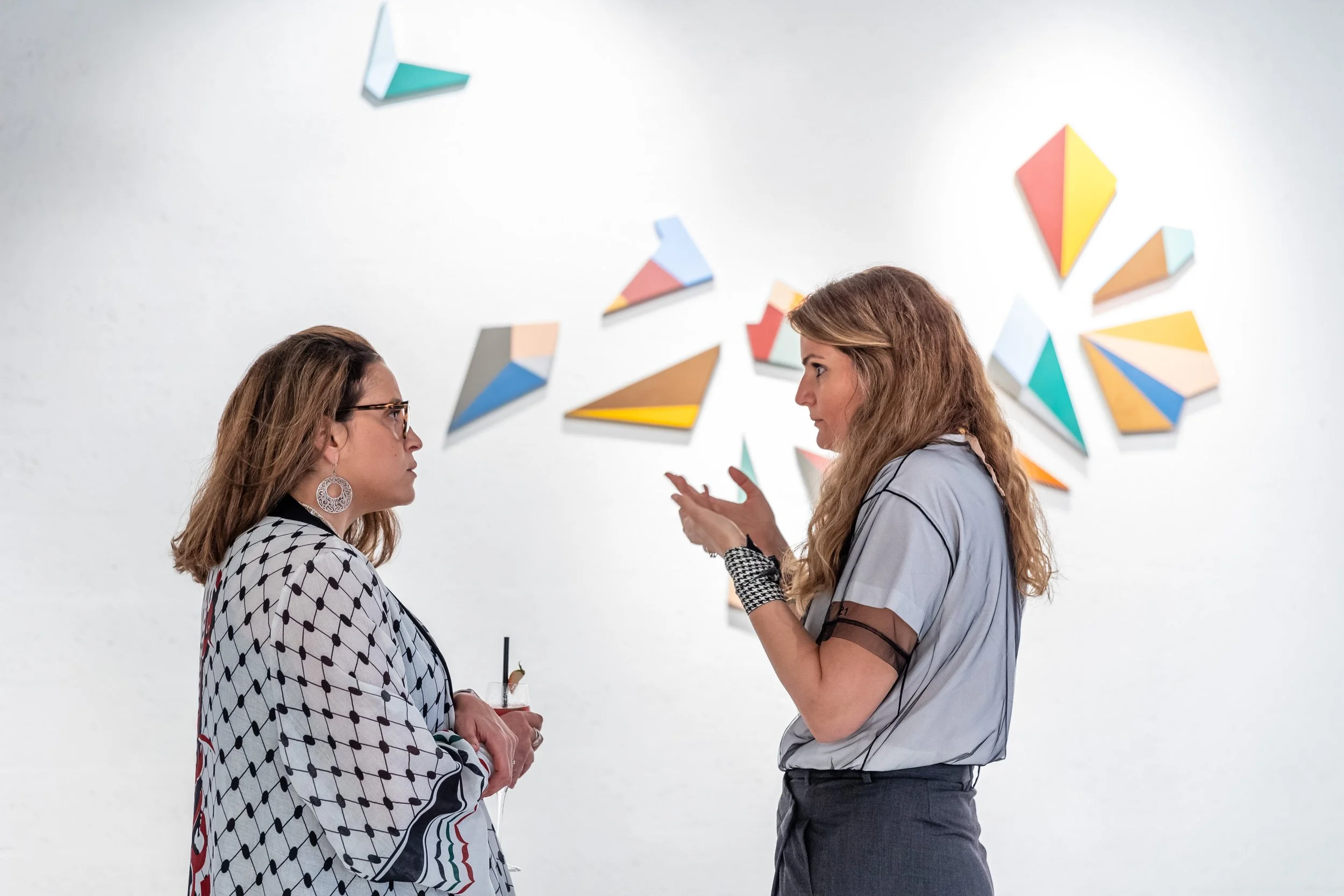 Two women in conversation at an art gallery, with colorful geometric artwork on the white wall behind them.