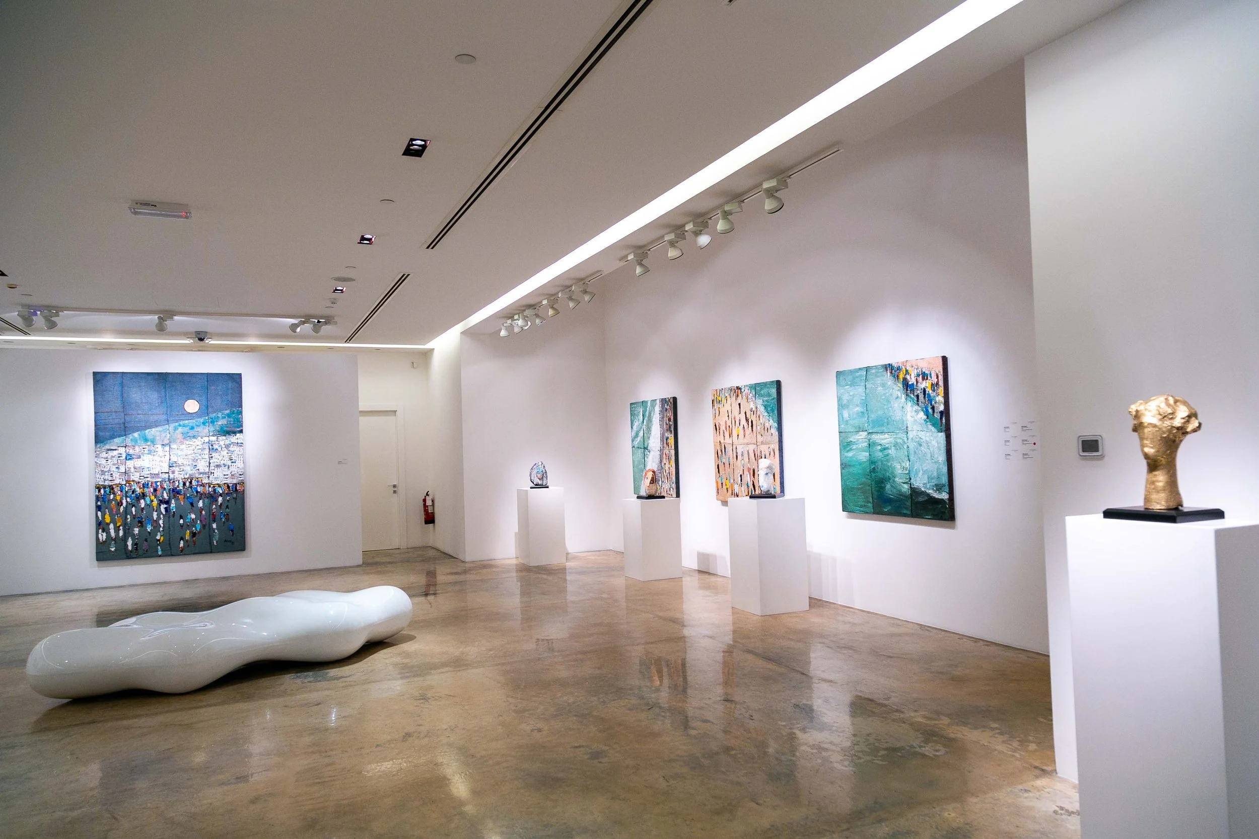 An art gallery with white walls and a polished concrete floor, featuring various abstract paintings on the walls and sculptures on pedestals, including a golden head sculpture and a modern, organic white bench in the middle.