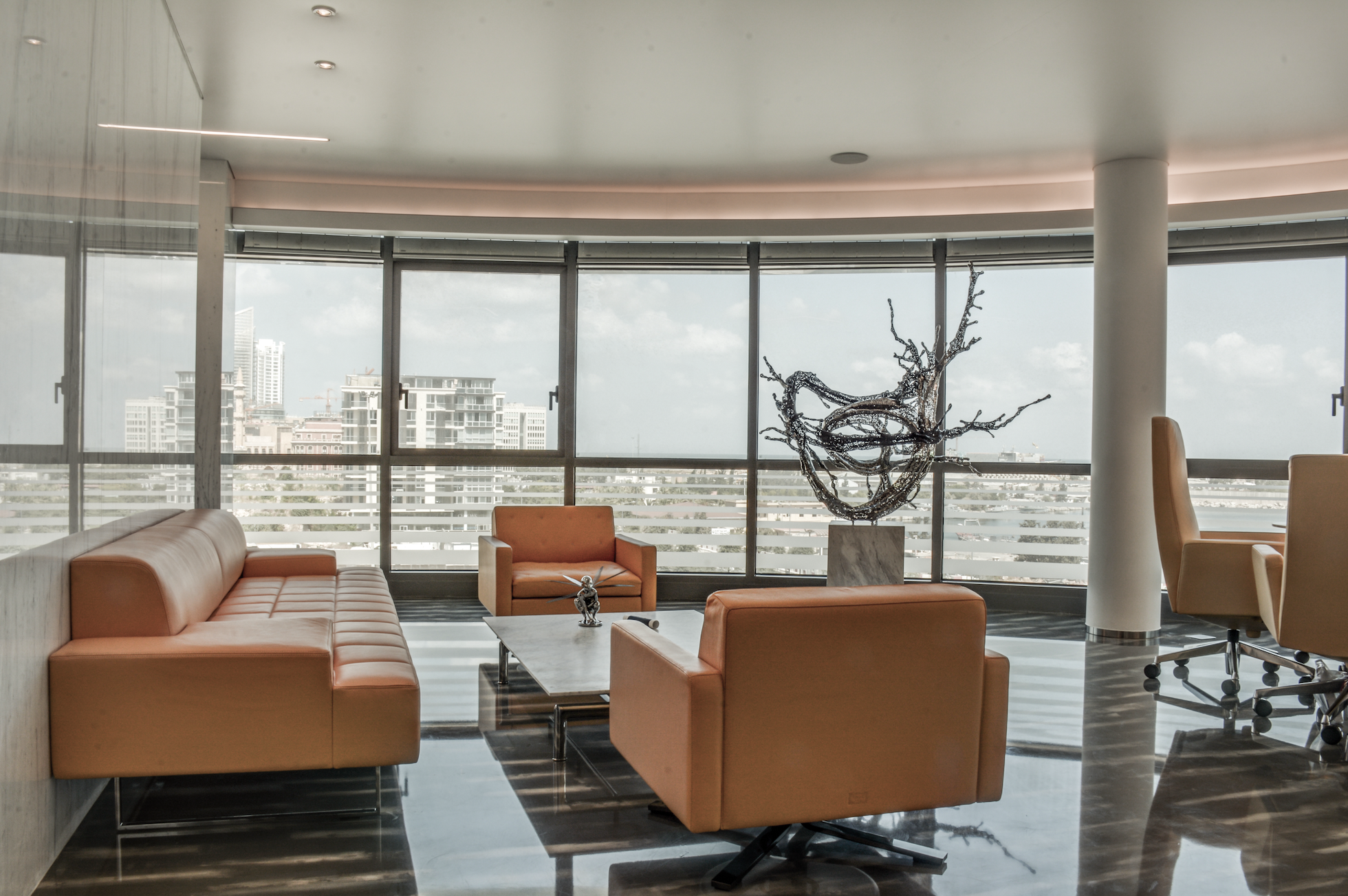 Modern office lounge area with orange leather sofas, a marble coffee table, a tall abstract sculpture, and floor-to-ceiling windows with city skyline view.