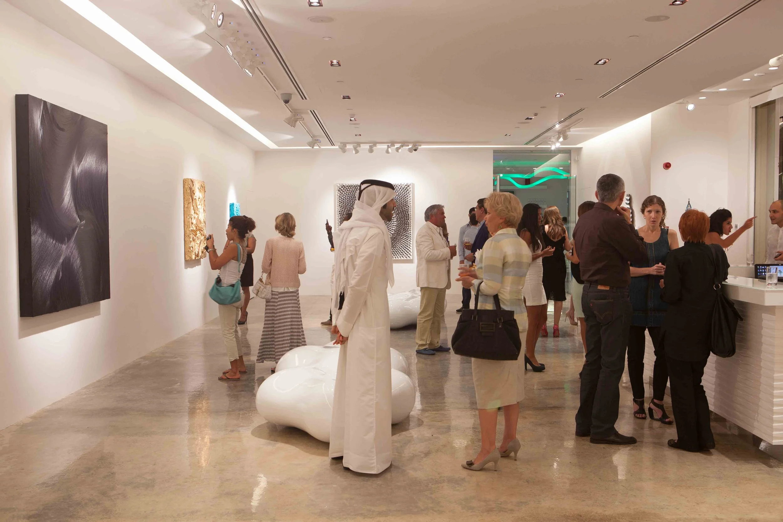 People gathered in an art gallery, observing and discussing artworks displayed on white walls, with some taking photos and others engaging in conversation, in a well-lit, modern space with artwork and sculptures.