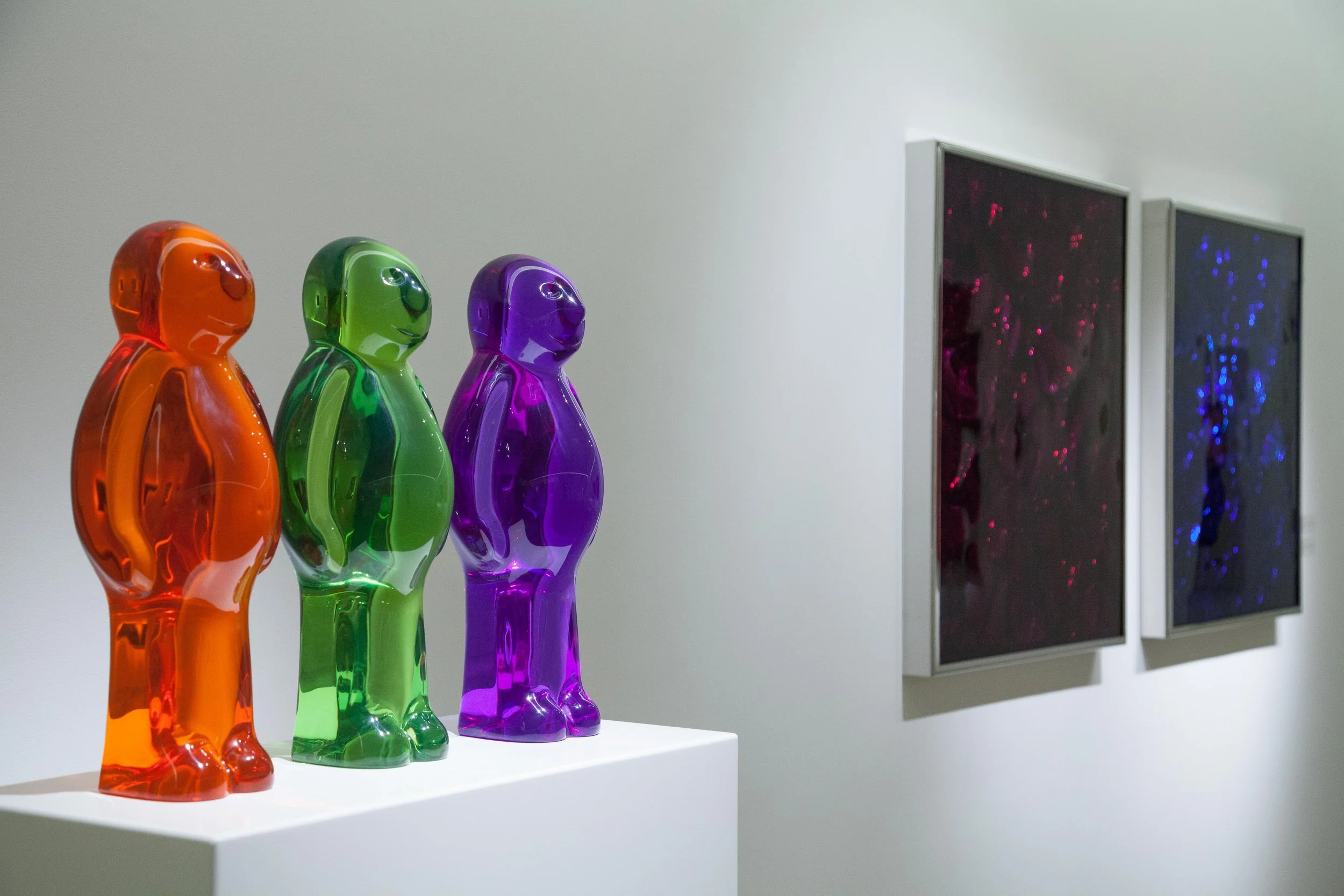 Colorful glass sculptures of three standing figures in orange, green, and purple, displayed on a white pedestal against a white wall, with two framed digital art pieces featuring abstract patterns in red and blue on the wall.