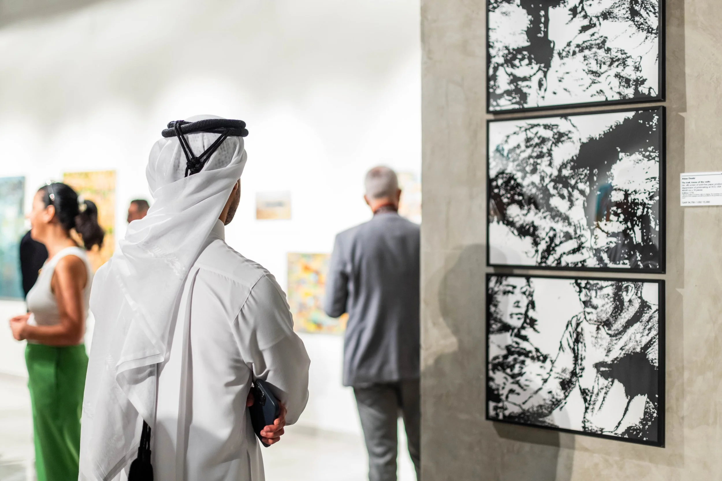 People viewing black-and-white artwork in an art gallery, with one man in traditional Middle Eastern attire and several other visitors.