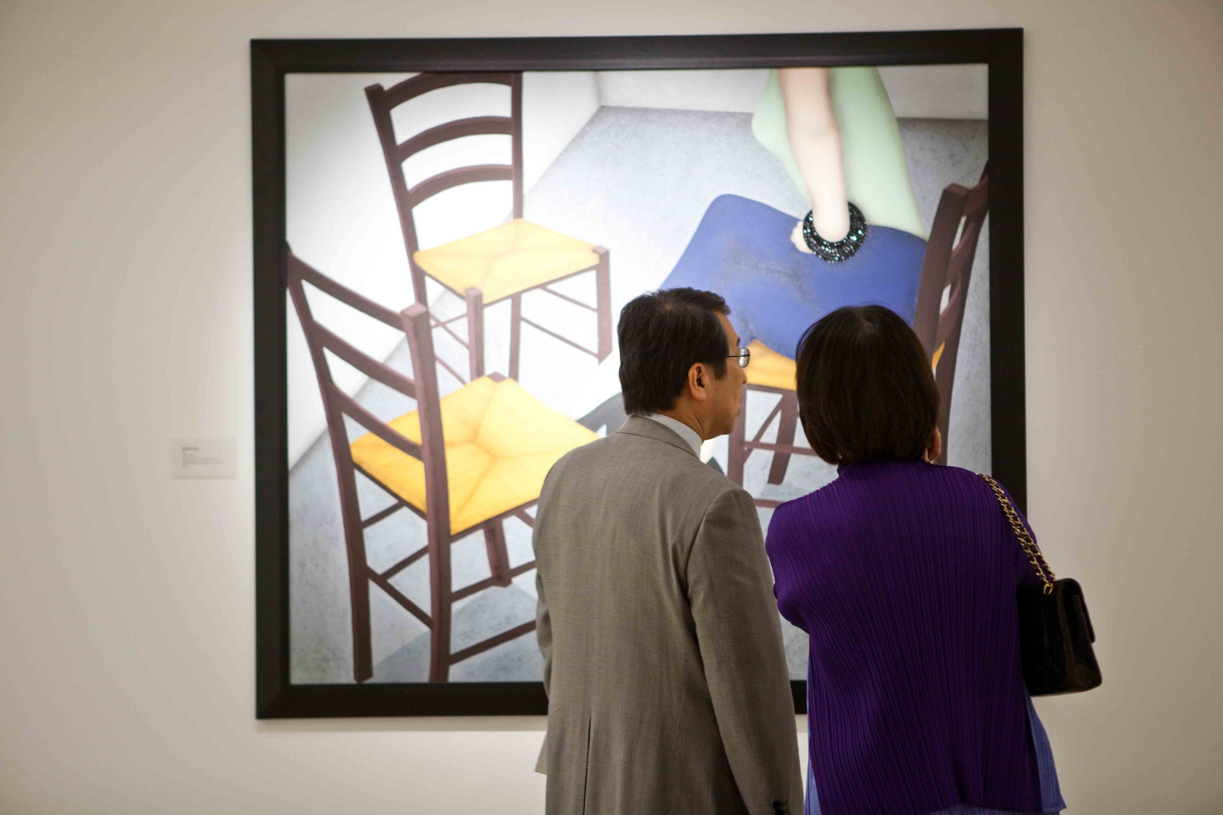 Two people standing in front of an artwork featuring four empty chairs, with a pair of legs visible in the background.