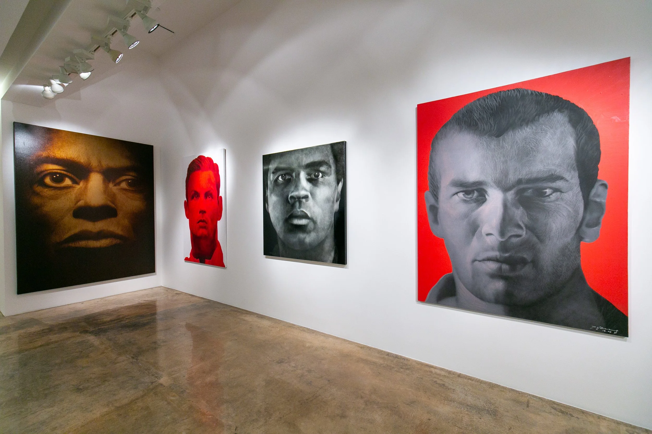 Gallery wall displays four large portrait paintings of diverse individuals, with detailed facial features, in black, red, and grayscale, inside an art gallery with white walls and track lighting.