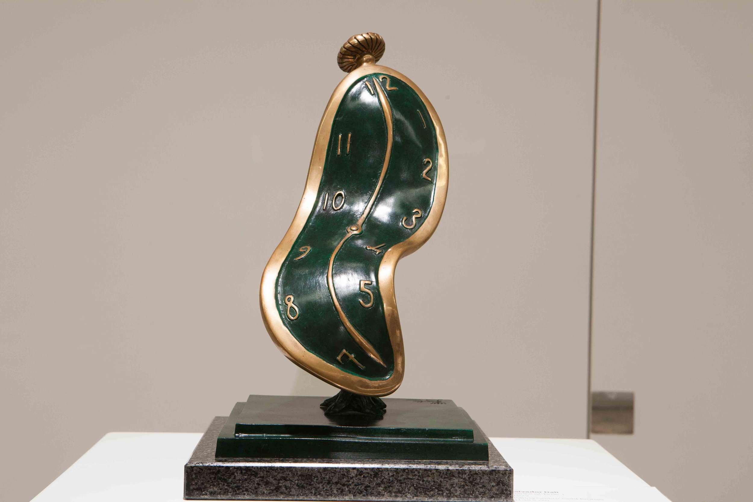 A surreal sculpture resembling a melting clock with a green face, gold edges, and numbers, mounted on a black base.