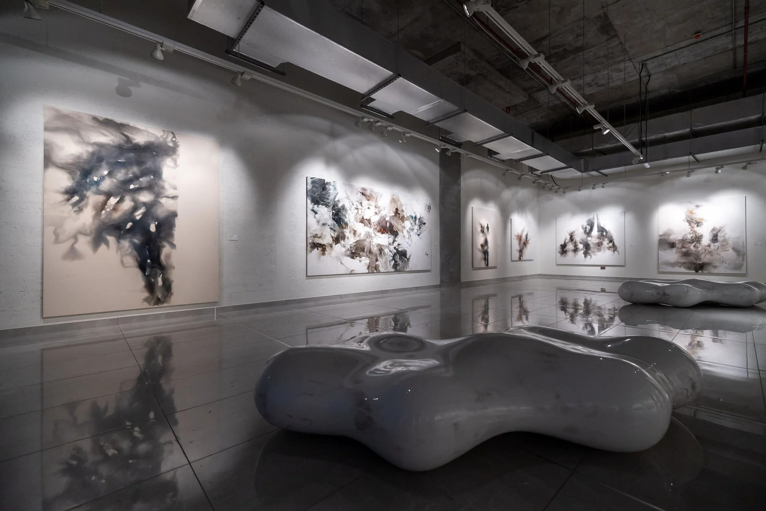 Modern art gallery featuring abstract paintings on white walls and glossy, organic-shaped benches on a reflective floor.