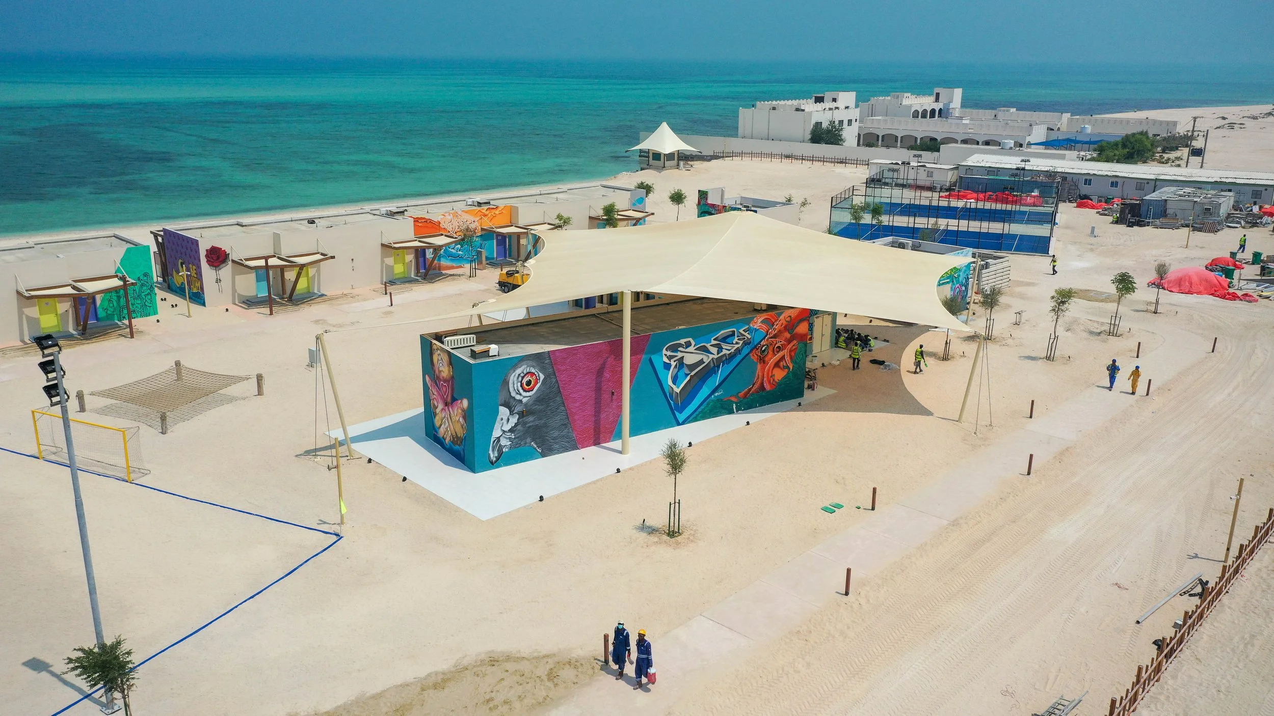 Construction site at a beach with buildings, colorful murals, and workers installing structures under a large canopy, near the ocean with sand and water.