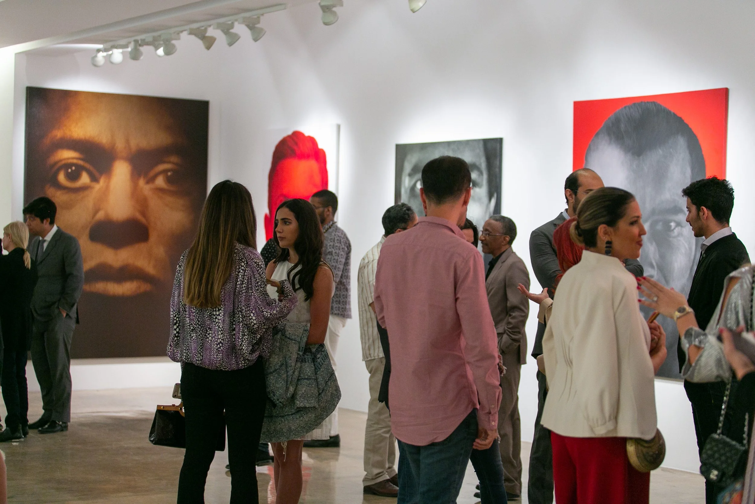 Gallery with people viewing large portrait artworks on white walls, artwork features close-ups of human faces in various colors.