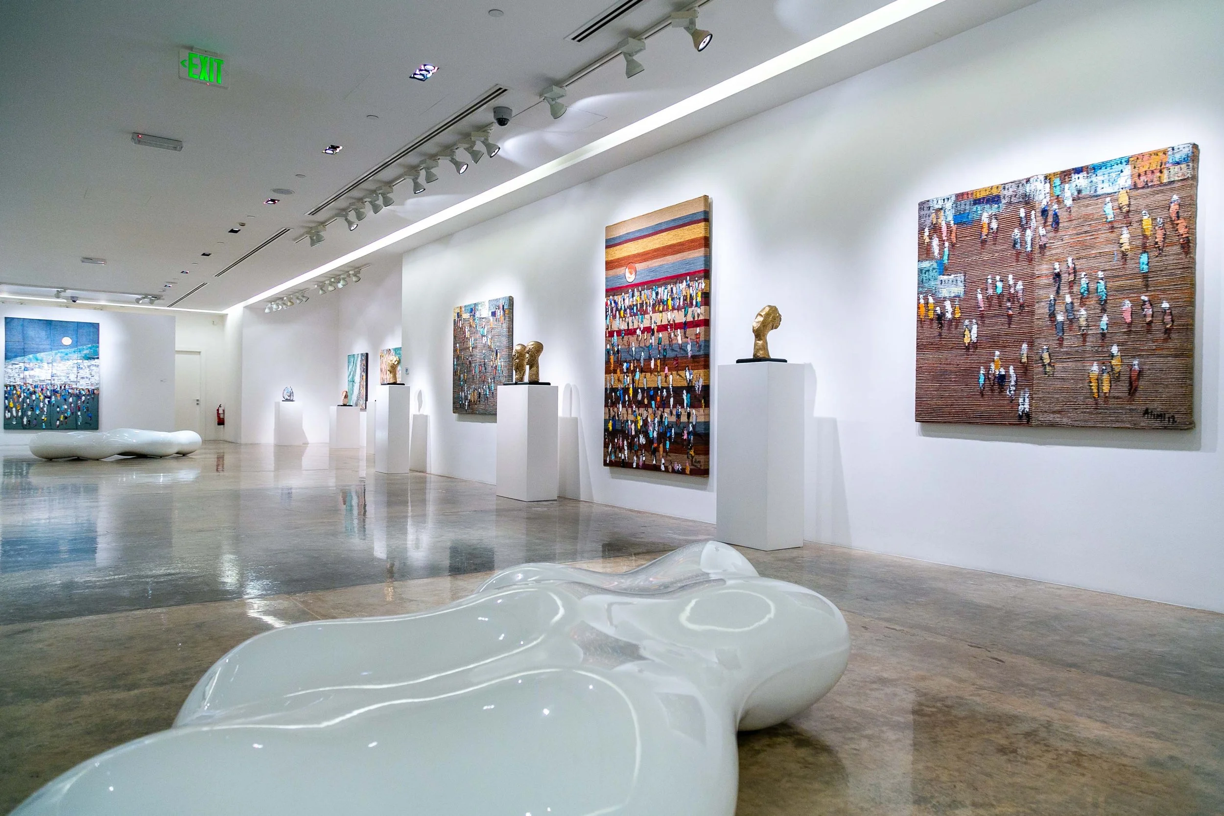An art gallery with several colorful paintings on white walls, sculptures on white pedestals, modern white benches, and polished concrete floors.