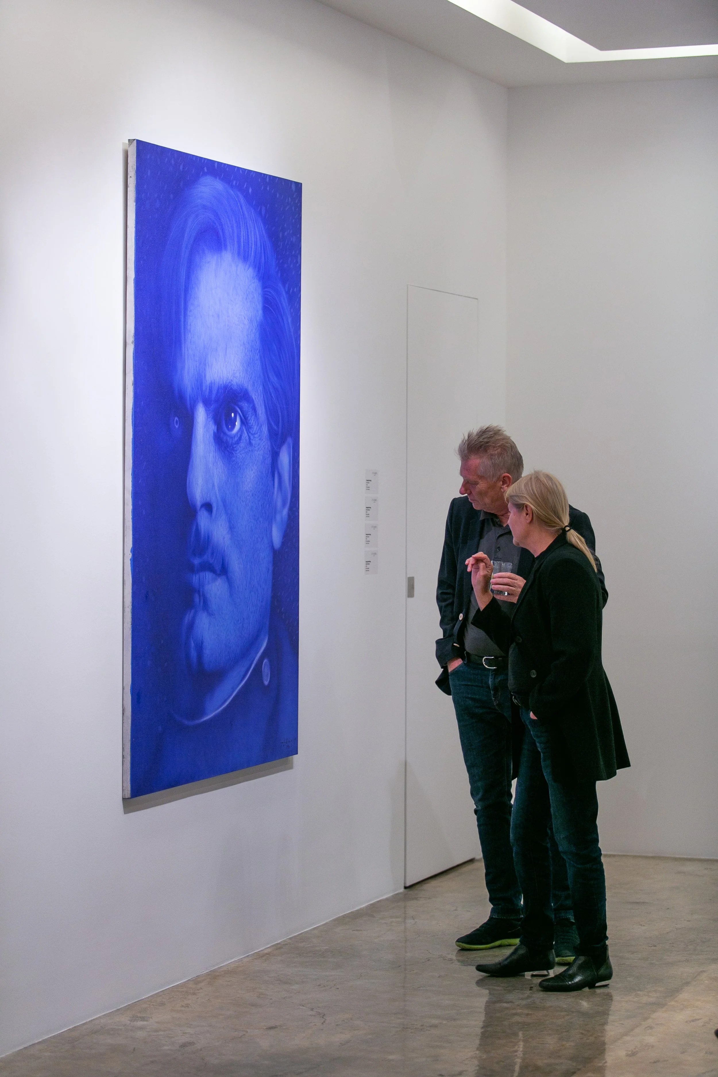 Two people, a man and a woman, are viewing a large blue monochrome portrait of a man with light-colored hair at an art gallery.