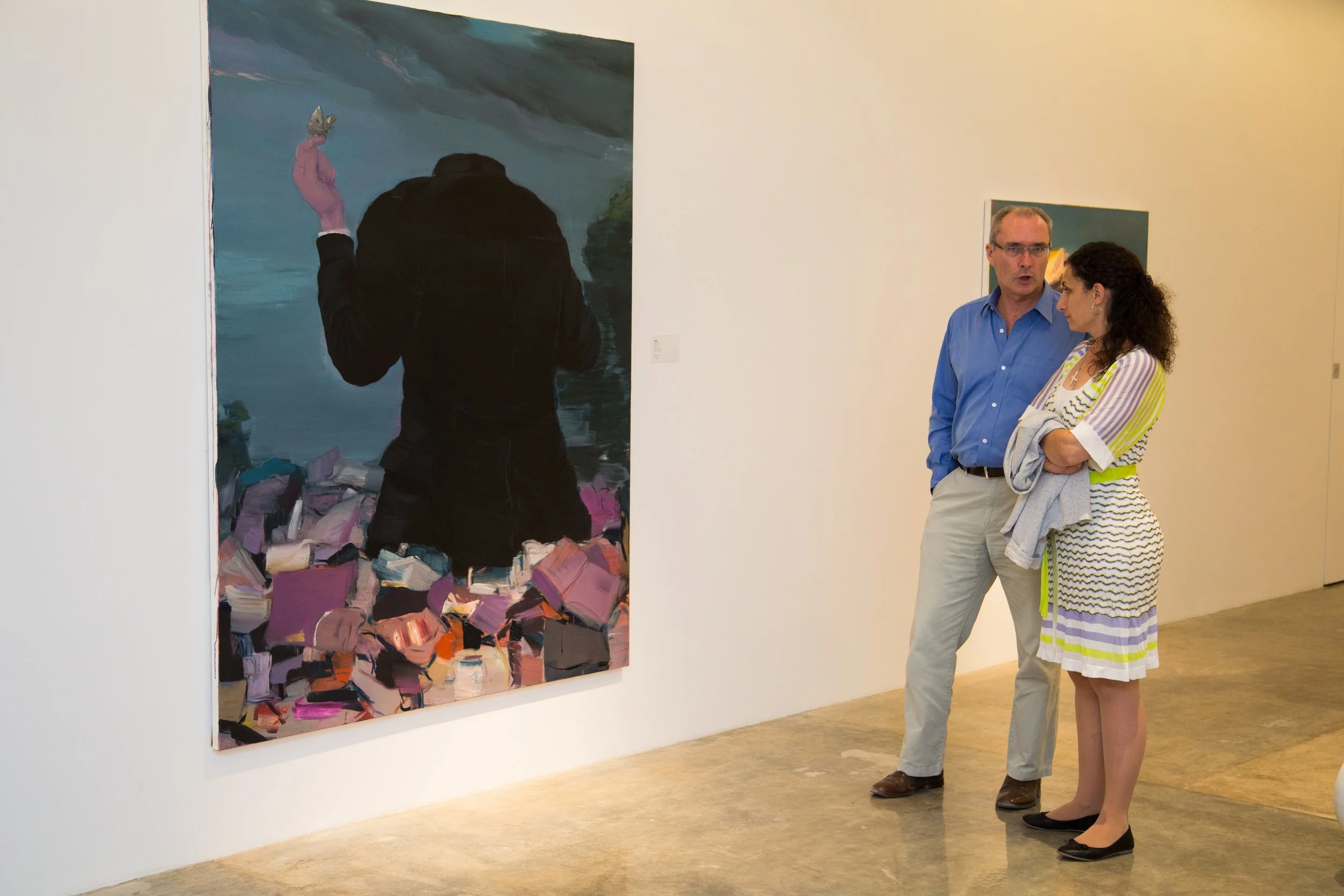 Two people, a man and a woman, are having a conversation in an art gallery next to a painting. The woman wears a striped dress, while the man wears a blue shirt and beige pants.