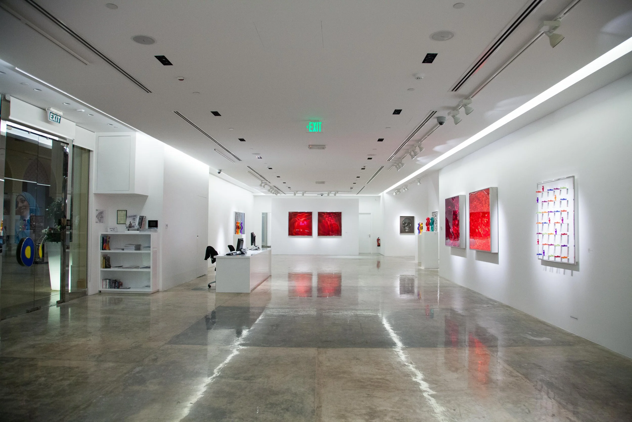 Interior view of an art gallery with white walls, featuring several abstract paintings and sculptures. The gallery has polished concrete floors and ceiling-mounted track lighting.