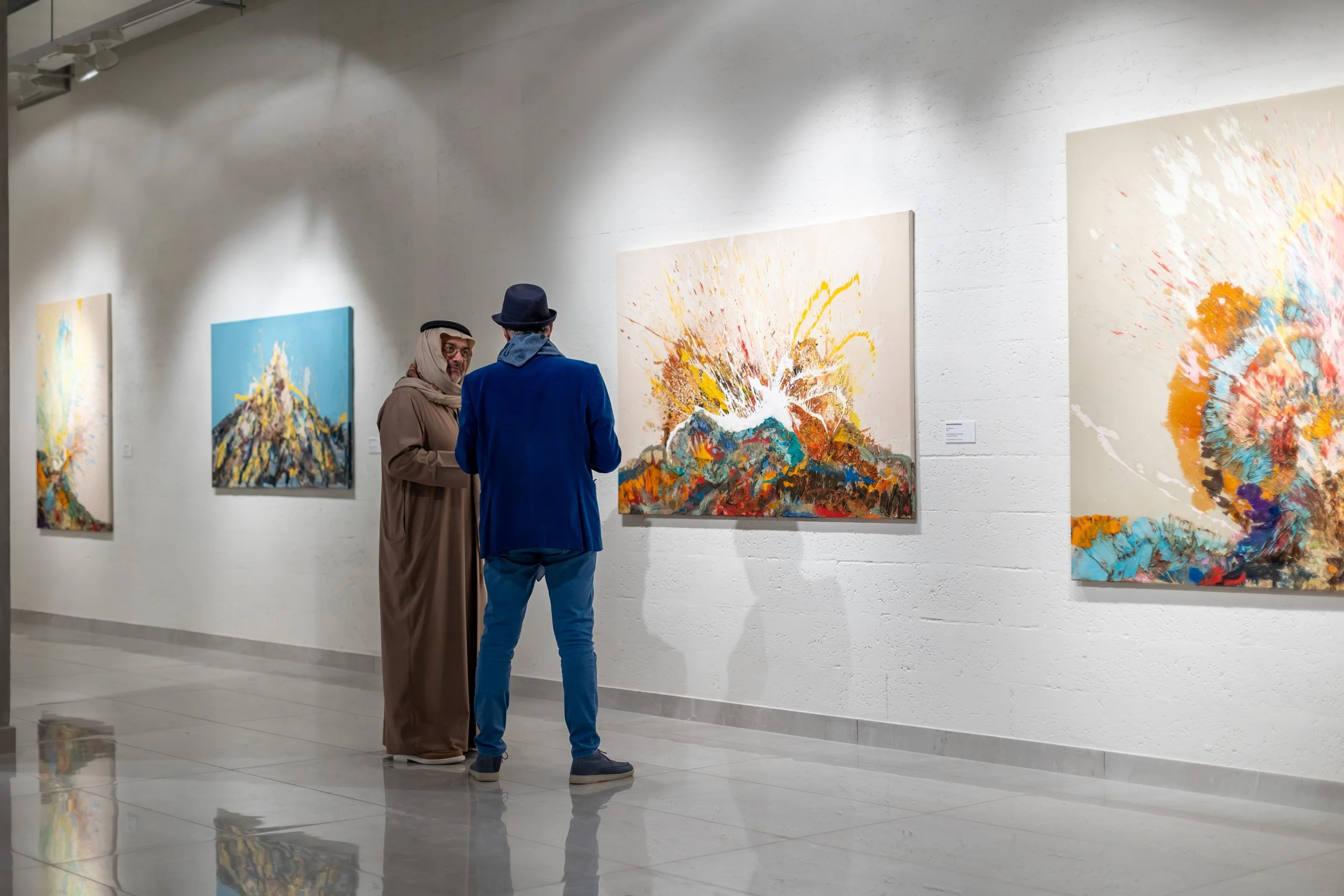 Two men in an art gallery, one wearing traditional Middle Eastern attire and the other in a blue jacket and hat, viewing colorful abstract paintings of explosions on white walls.