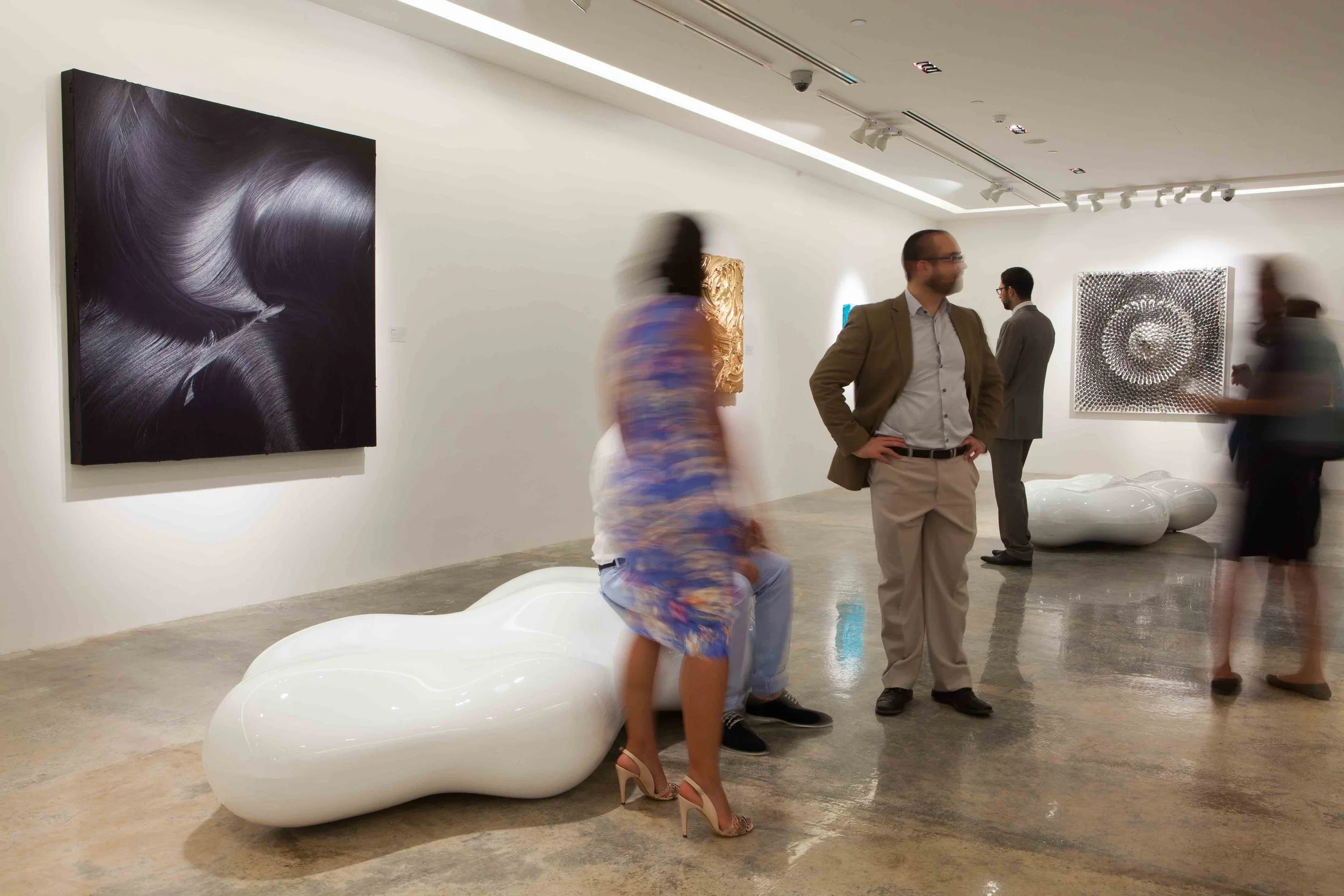 People viewing artwork in a modern art gallery. Art includes a large black and white abstract painting, a textured gold sculpture, a small blue piece, and a patterned black and white piece. There is also a white sculptural bench.