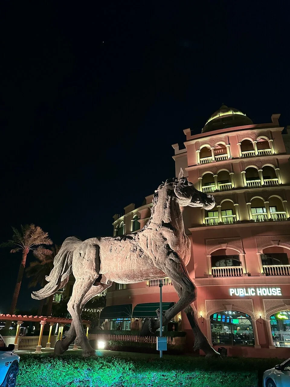 Large bronze horse statue in front of a pink, multi-story building with columns and balconies, nighttime scene with colorful lights illuminating the area.