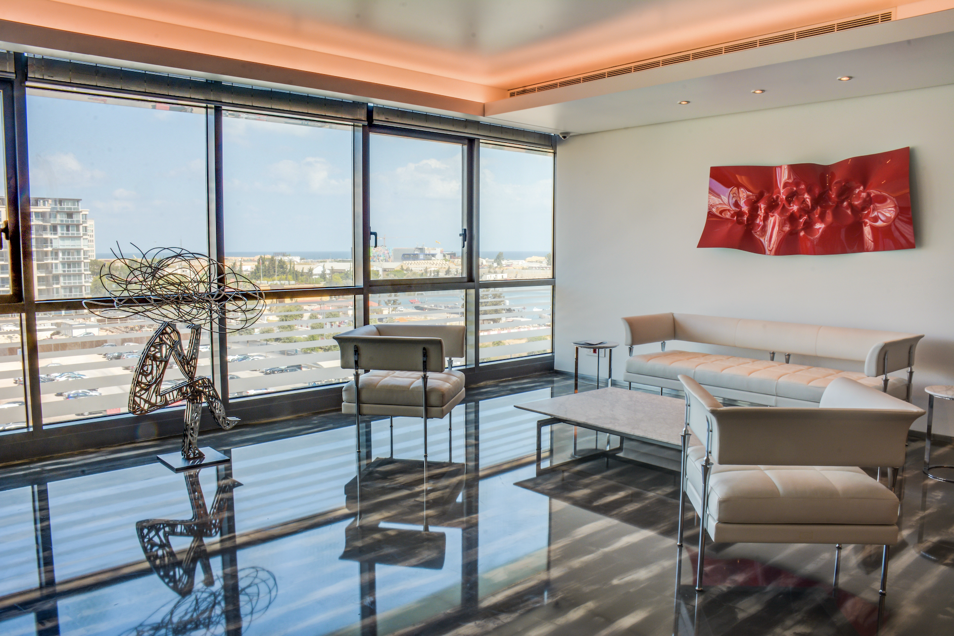Modern hotel lobby with large windows, contemporary white furniture, a red abstract wall art piece, a metal sculpture, and a view of the city and ocean outside.