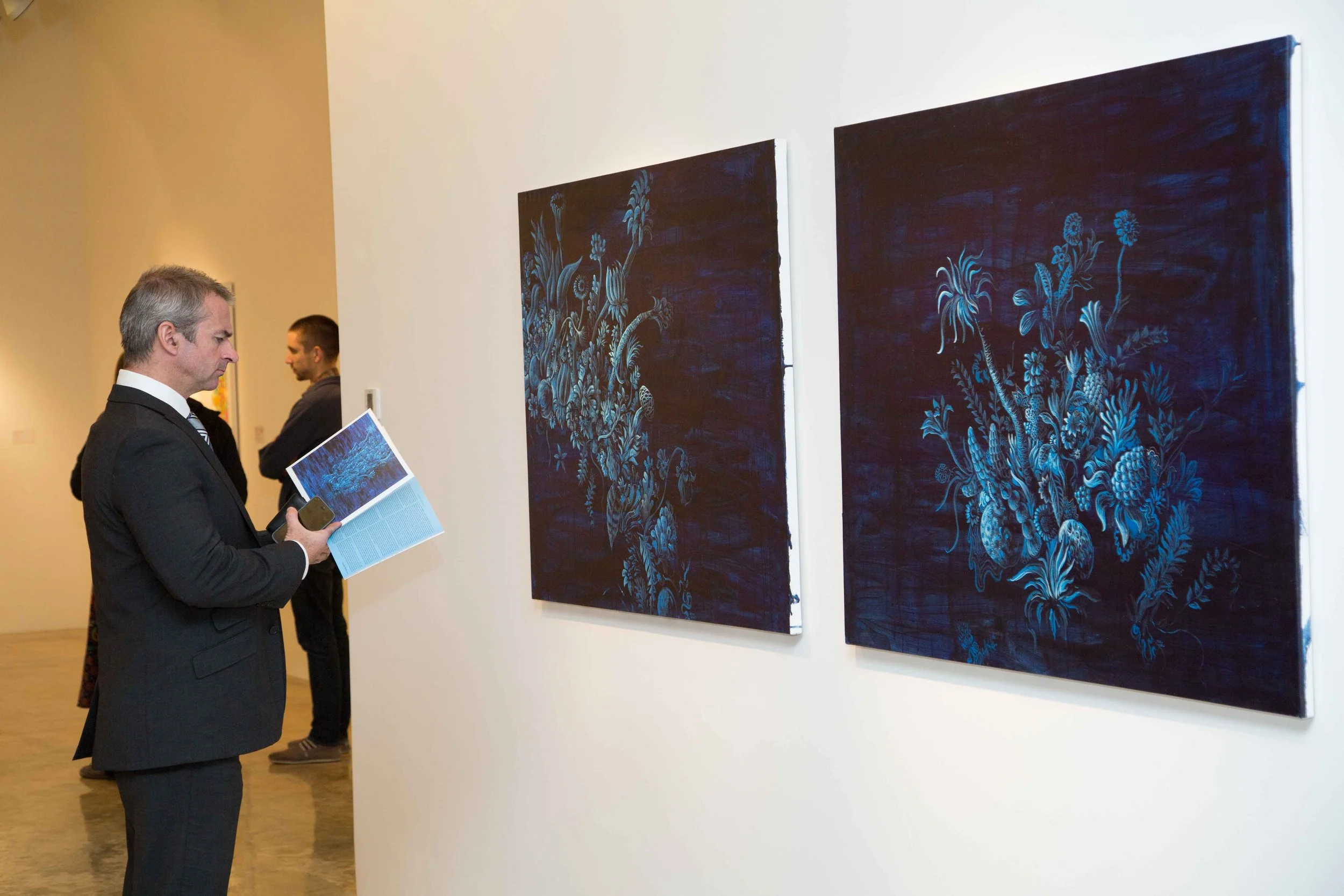 A man in a suit looking at two blue floral paintings on a white gallery wall, holding an exhibition catalog and a smartphone, with other visitors in the background.