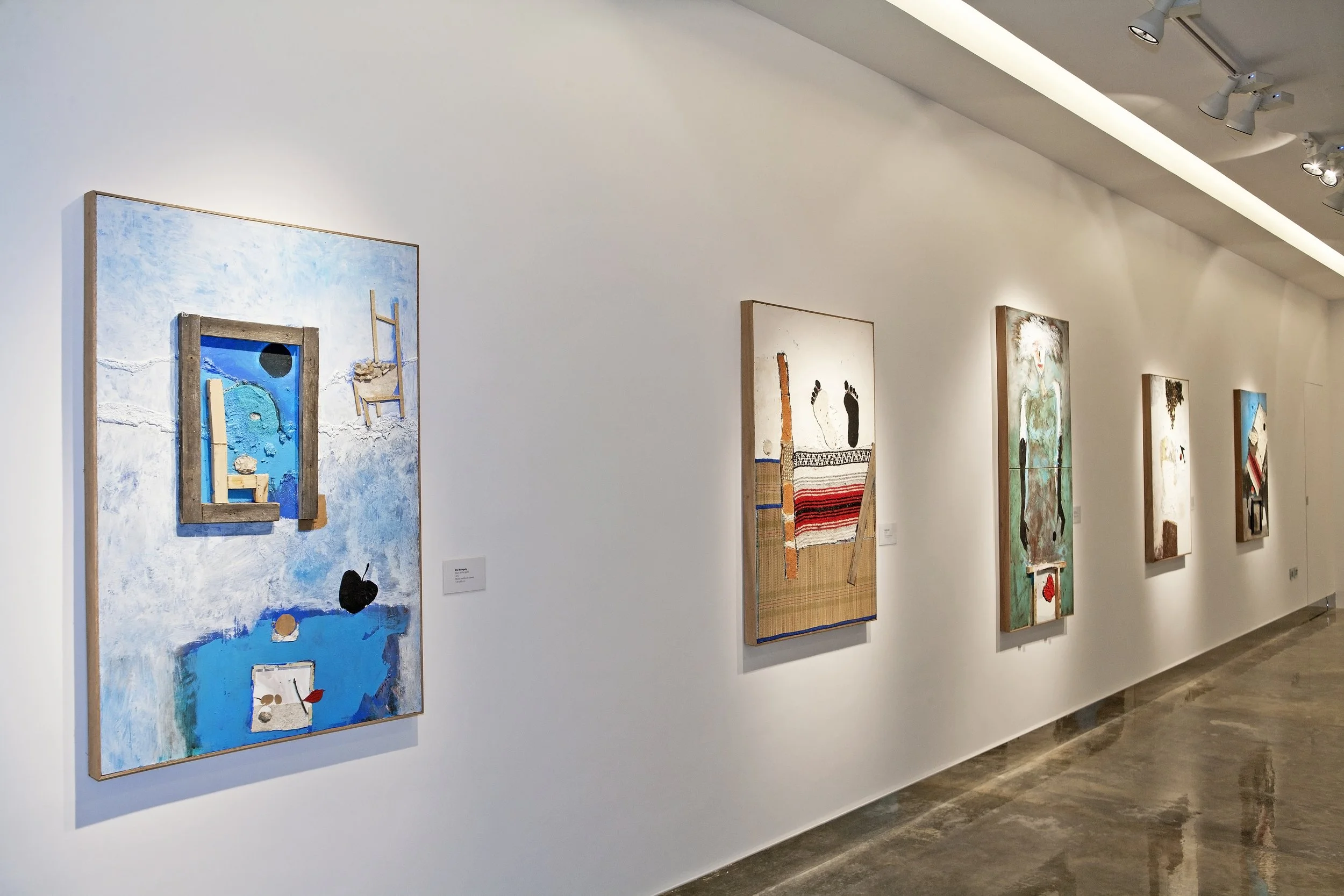 Gallery wall with multiple abstract paintings in a modern art museum. The paintings feature various textures, colors, and mixed media elements. The room has white walls and polished concrete floors with track lighting on the ceiling.